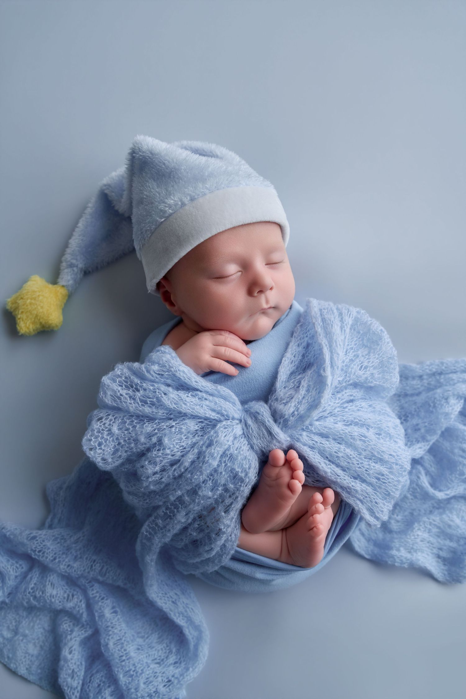 Sleeping newborn baby wrapped in soft blue knit blanket wearing a blue pointed hat with yellow star tassel.