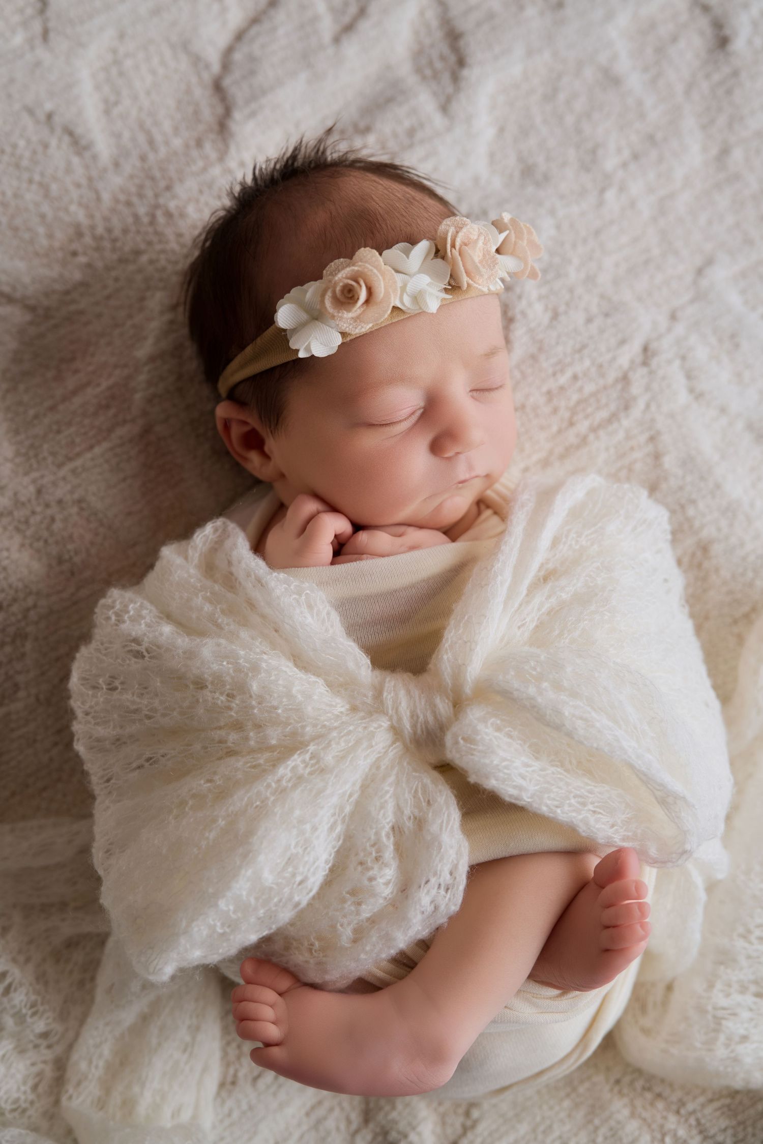 Sleeping newborn baby girl wrapped in white lace swaddle with floral headband on soft cream blanket.