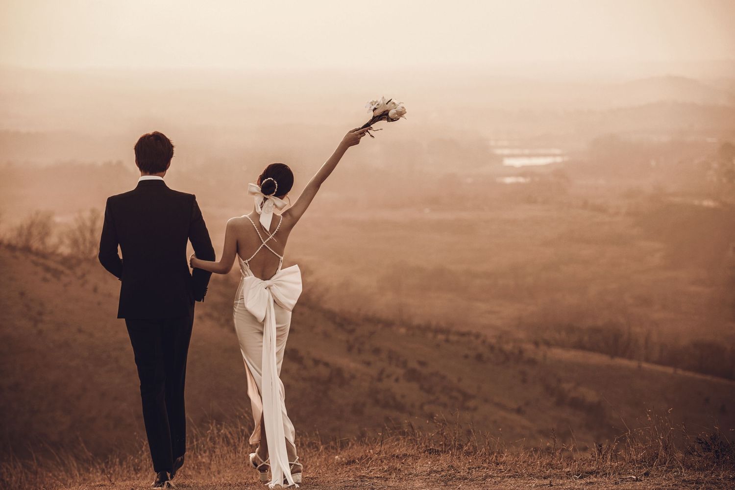 Bride in elegant backless white gown raises bouquet while walking with groom overlooking scenic countryside landscape.