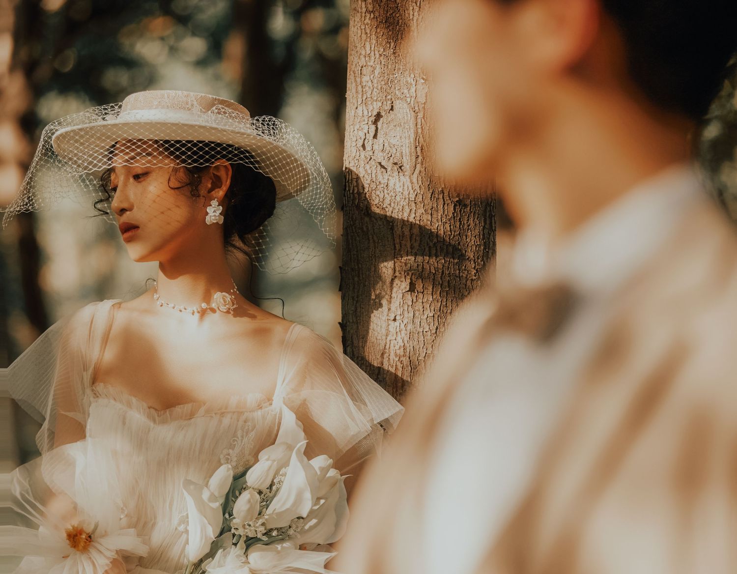 Elegant bride in hat and veil holding bouquet by tree.