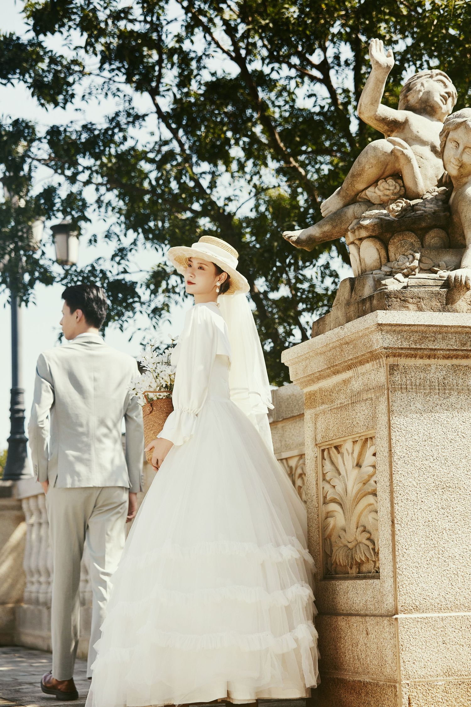 Bride in layered white gown and straw hat stands near ornate stone sculpture with groom in gray suit.