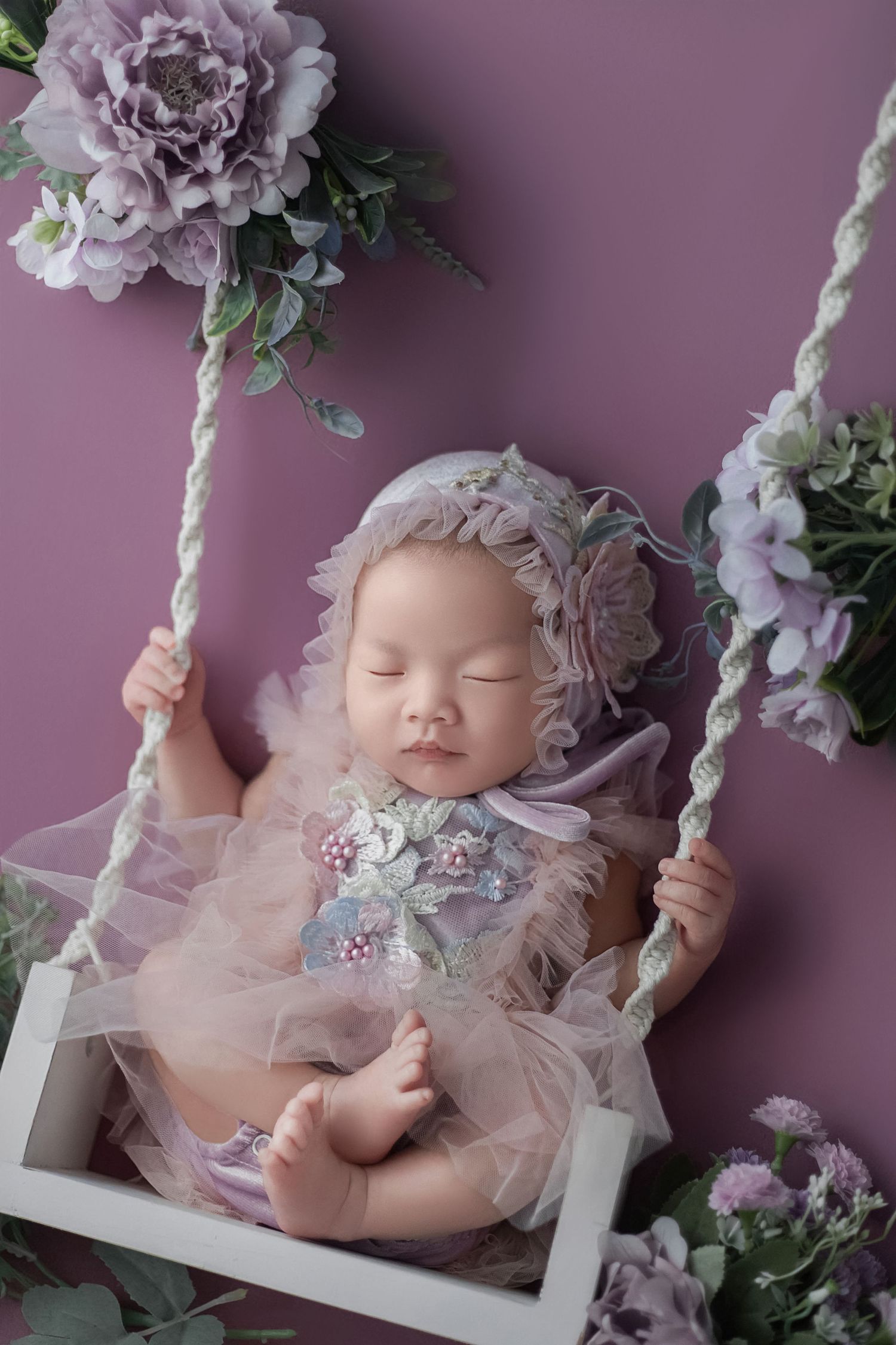 Sleeping newborn baby girl in purple floral outfit on a white swing surrounded by lavender flowers.