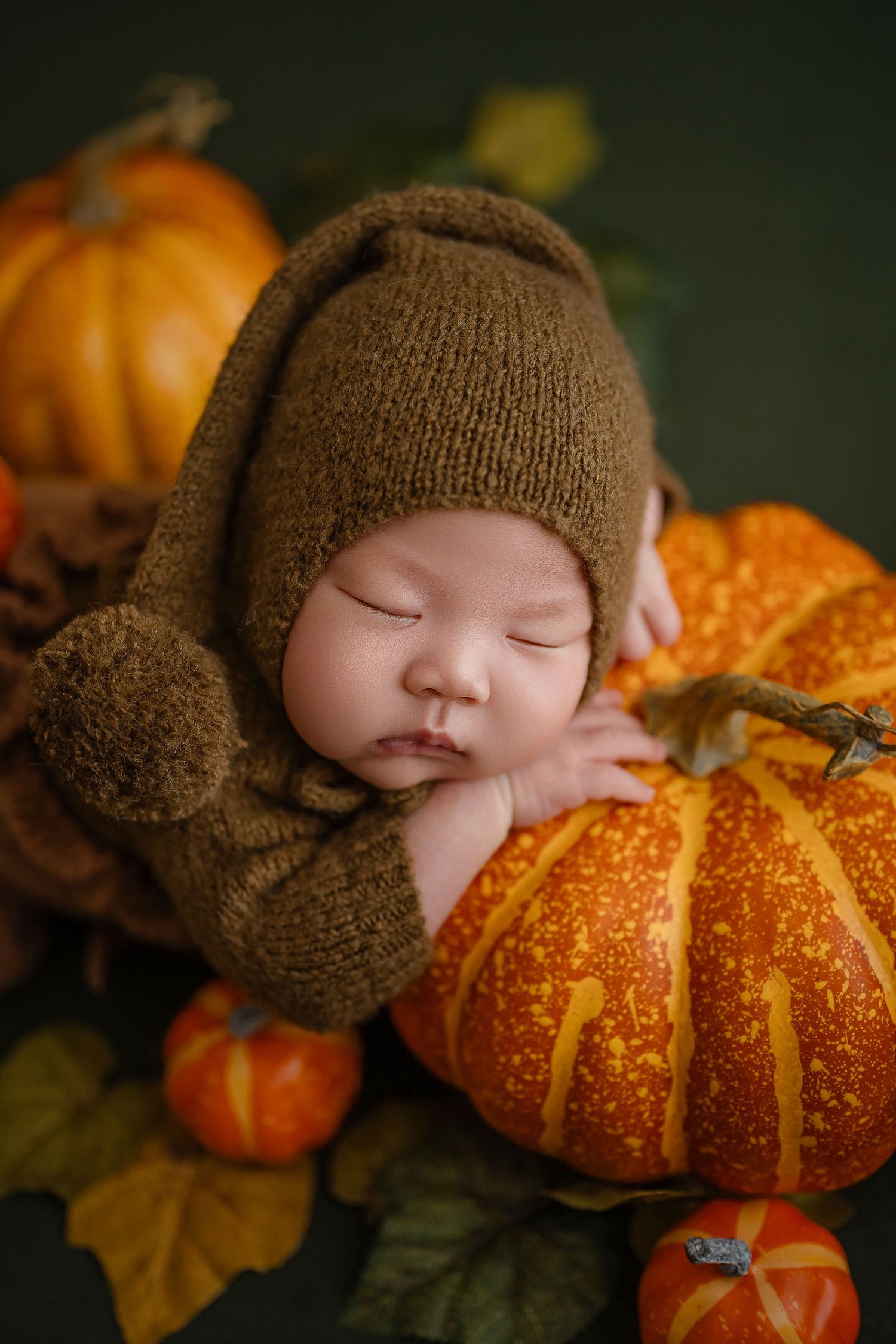 Newborn baby in brown knit hat sleeps peacefully resting on a large orange pumpkin surrounded by autumn decor.