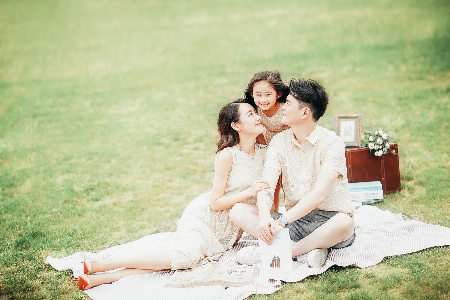 Loving couple with child sharing a kiss on a picnic blanket on a lush green grass field.