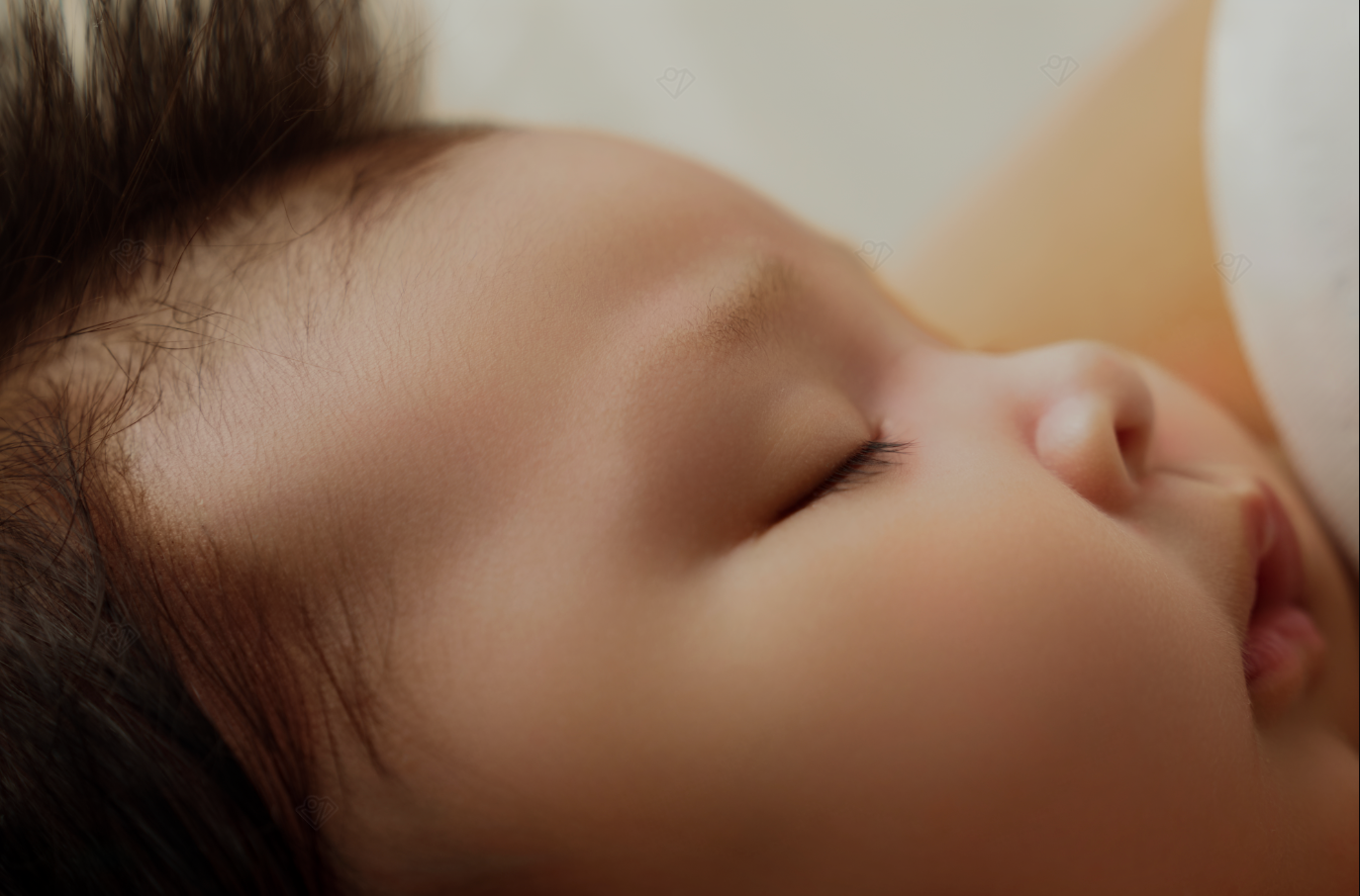 Close-up of a sleeping newborn baby with dark hair, eyes closed, resting peacefully against a soft background.