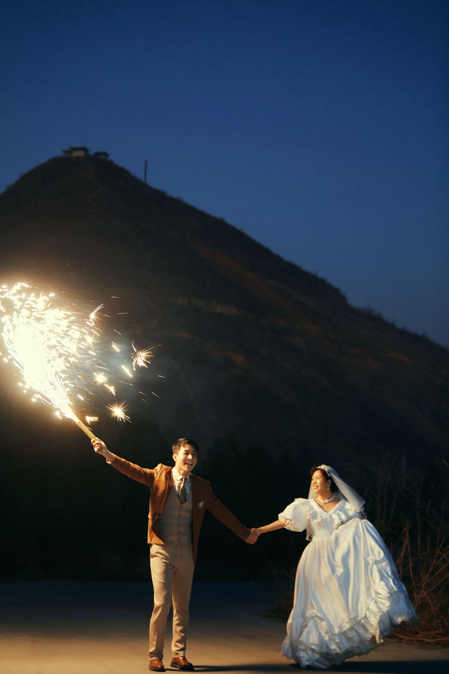 Newlywed couple holding sparklers at night with mountain silhouette in background during wedding shoot.