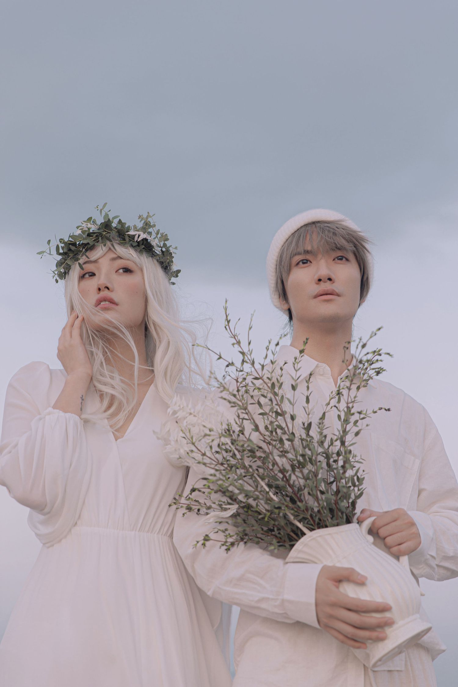 Young couple in white outfits stand against cloudy sky, man holding wildflowers, woman wearing leaf crown.