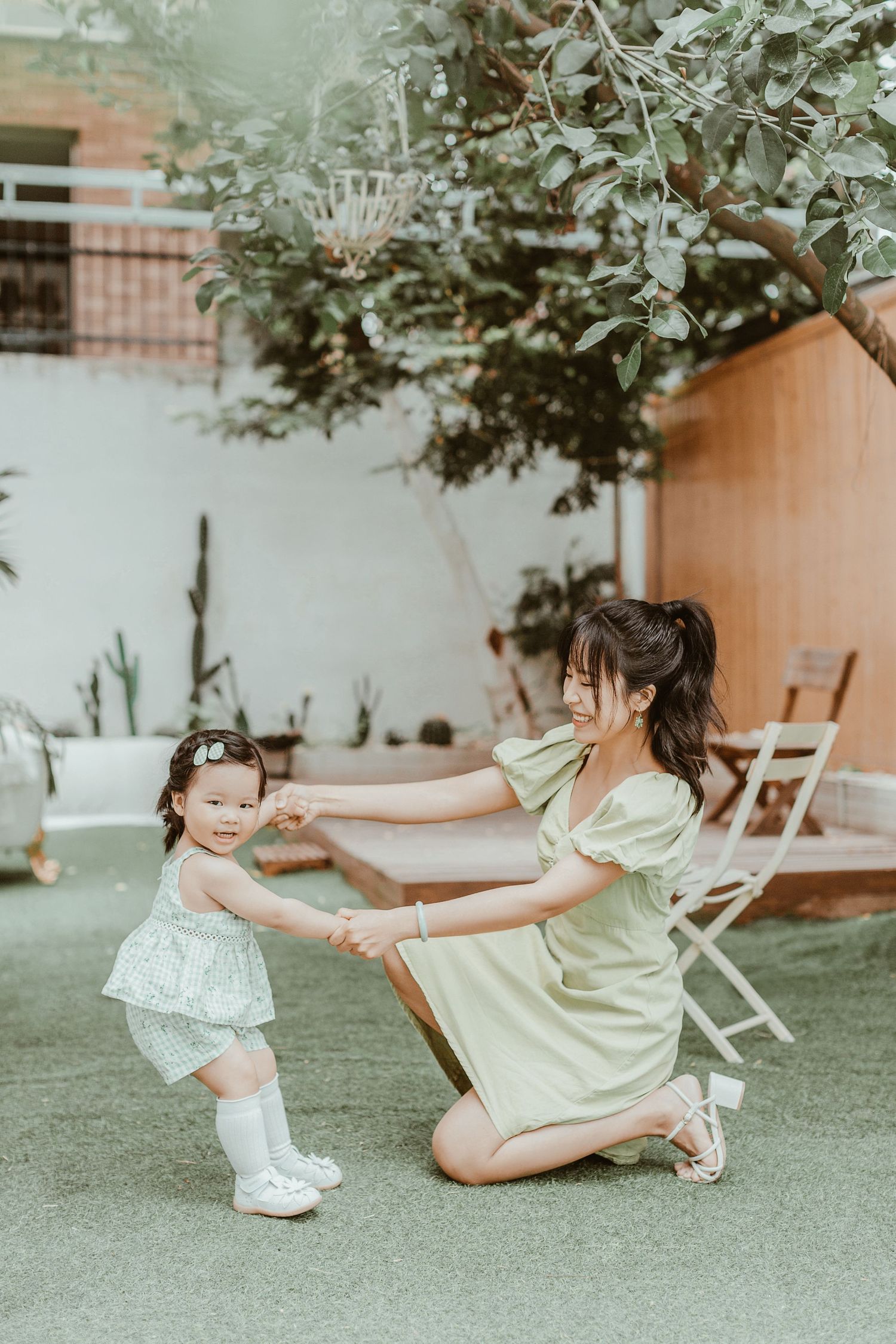 Smiling Asian mother in green dress kneels to hold hands with her cheerful toddler daughter outdoors.