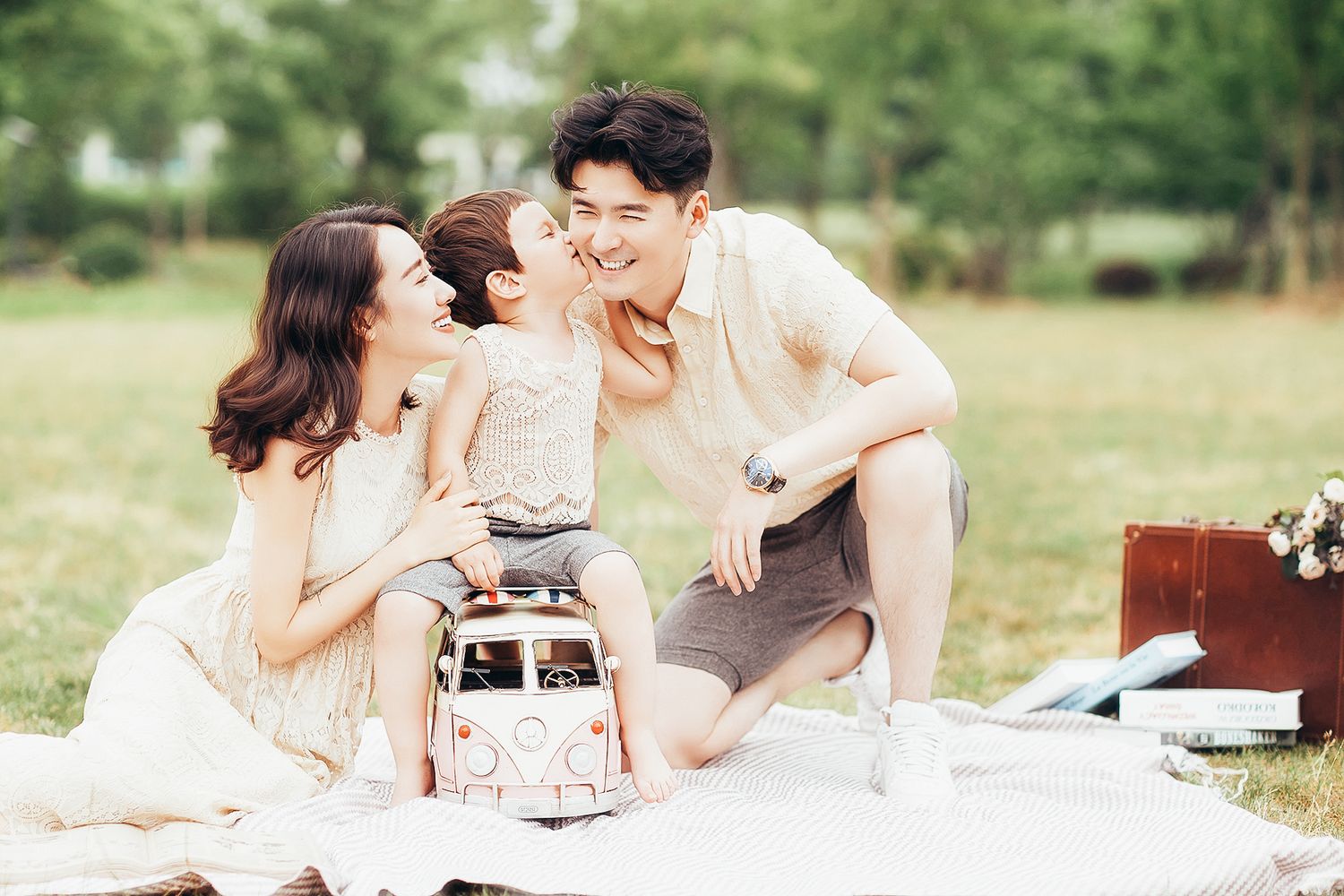 Smiling family of four sharing a joyful moment on a picnic blanket with a toy bus outdoors.