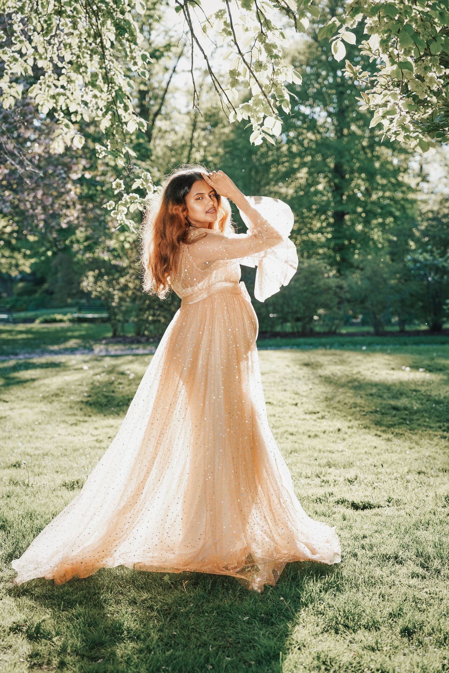 Woman in elegant champagne tulle gown poses gracefully in sunlit park surrounded by blooming trees and lush green grass.