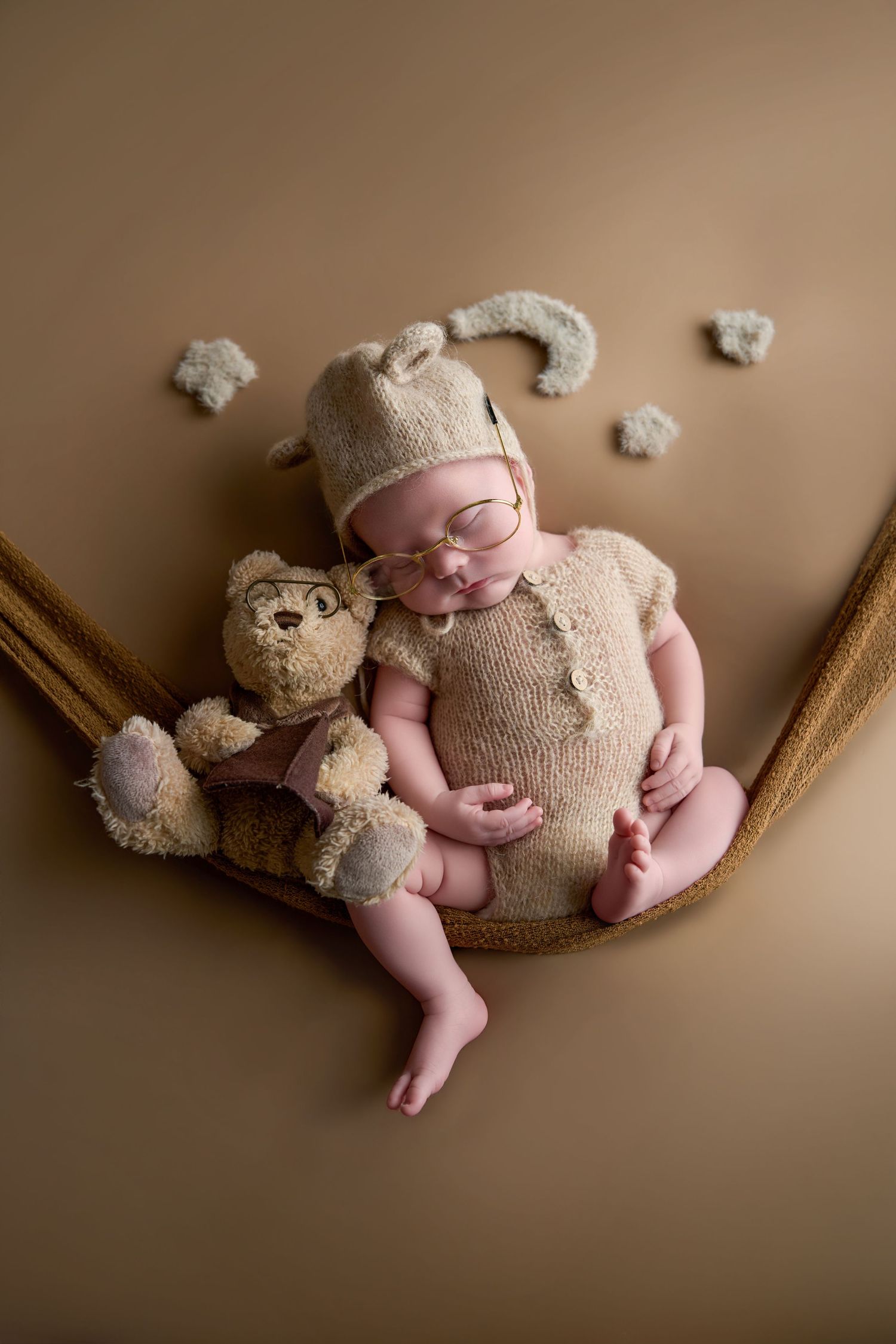 Newborn baby sleeping in a hammock wearing a bear knit outfit and glasses, cuddling a teddy bear on a beige background.