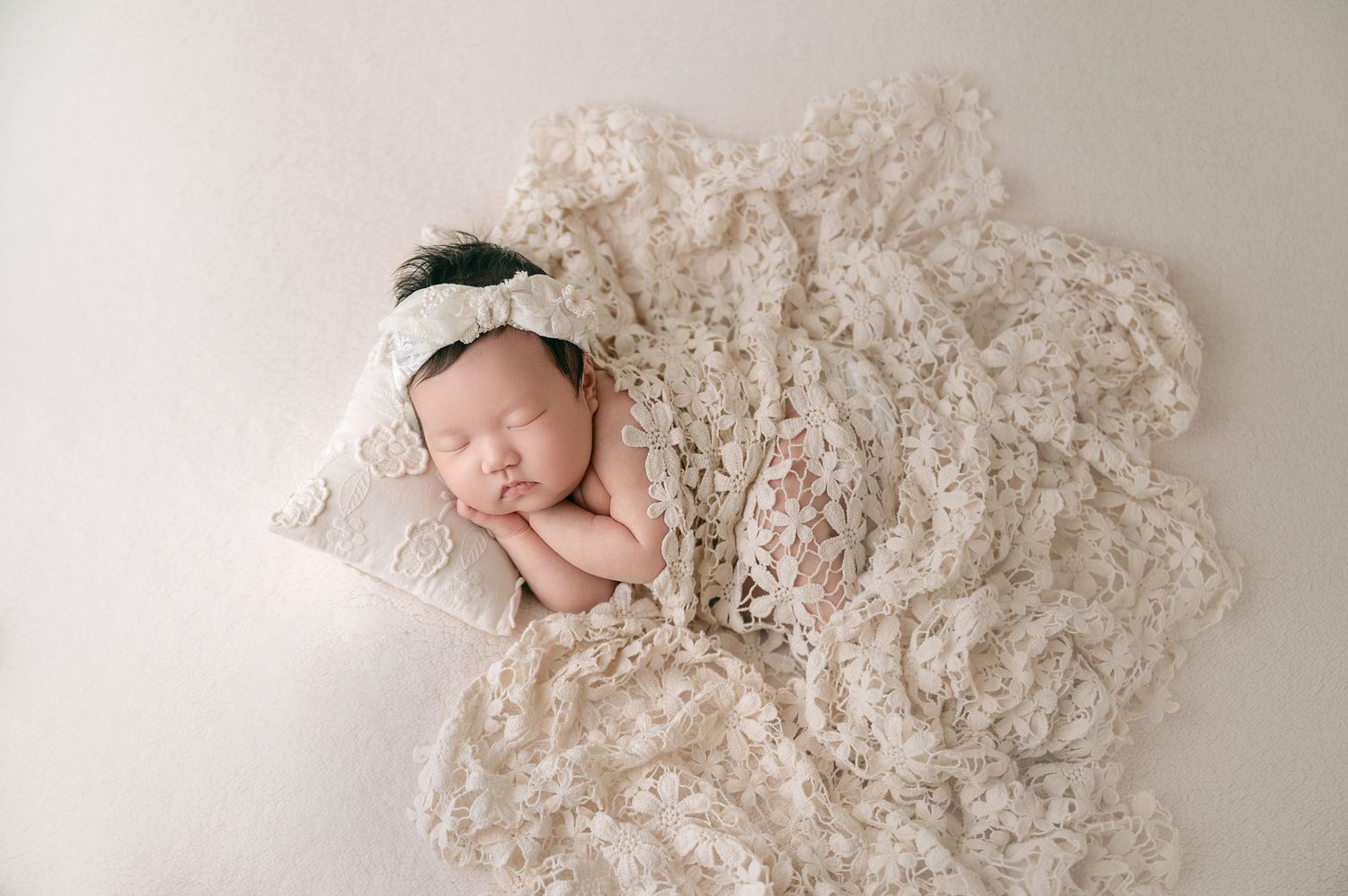 Sleeping newborn baby wrapped in cream lace blanket with bow headband resting on a lace pillow.