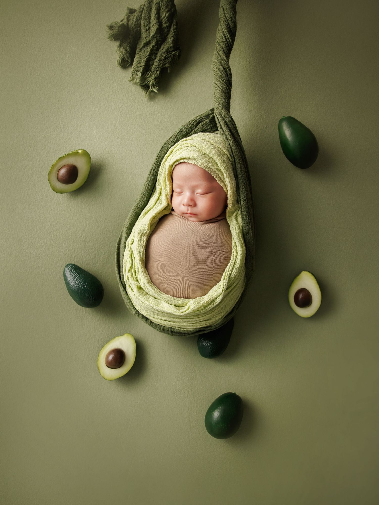 Sleeping newborn baby in avocado-shaped hanging prop with green wrap surrounded by avocados.