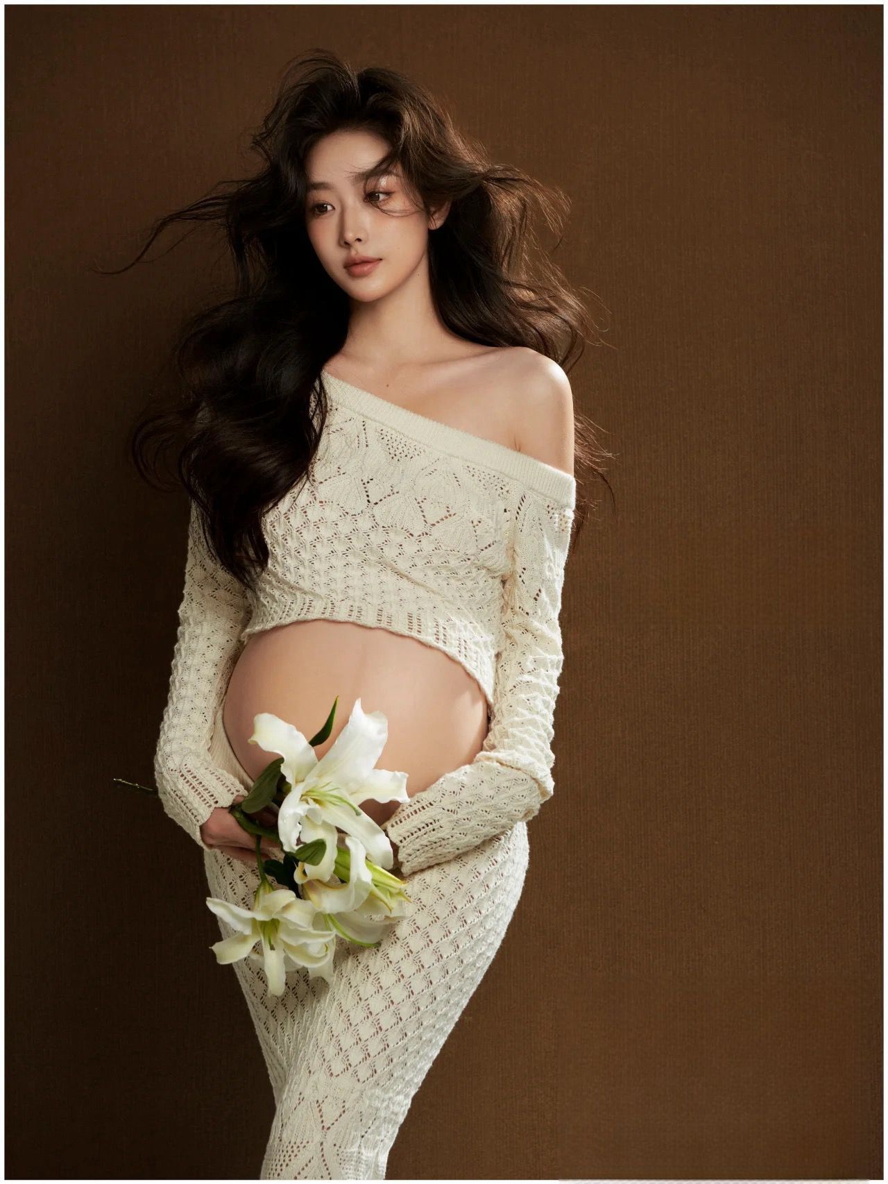 Pregnant model in cream off-shoulder cable-knit two-piece holding white lilies against brown background.