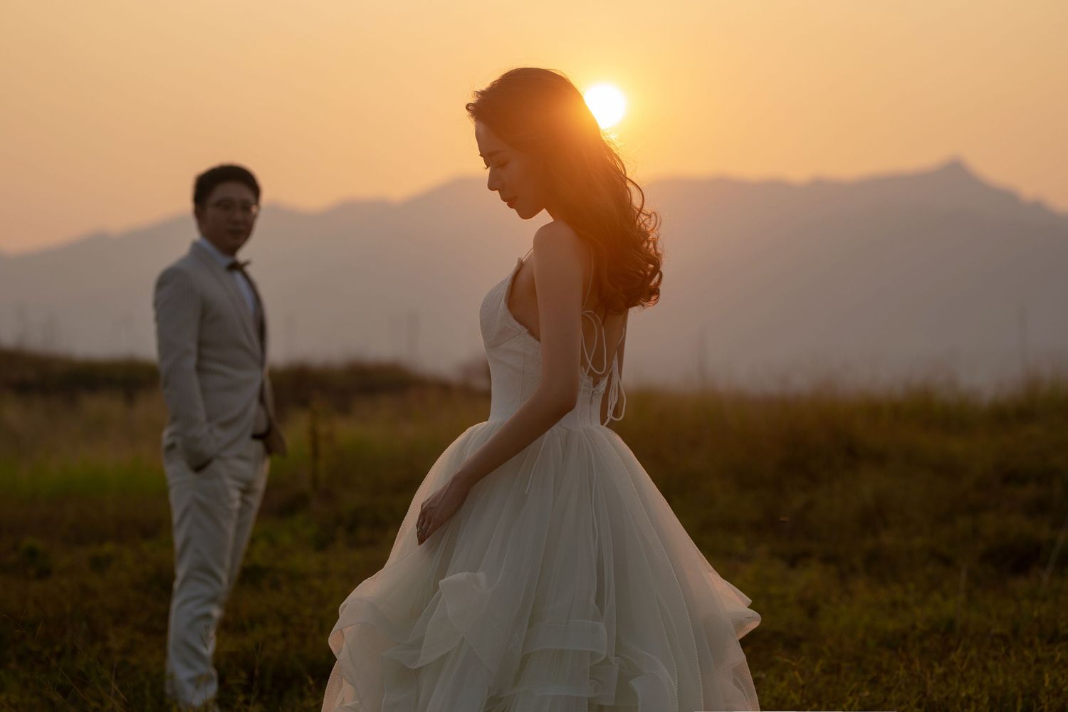 Bride stands gracefully in layered white gown as groom watches from behind during a golden hour mountain sunset shoot.