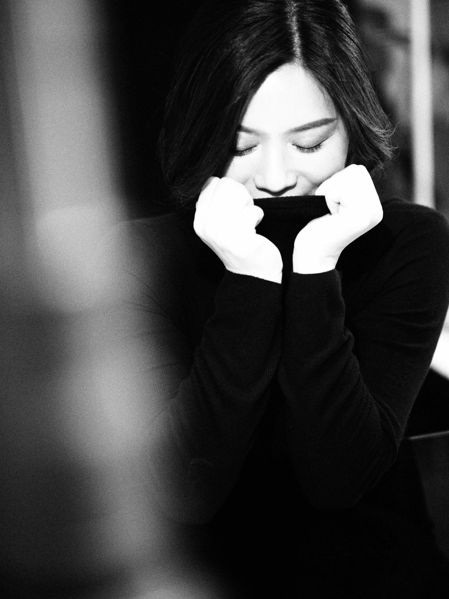 Black and white portrait of a woman in turtleneck sweater with hands raised to face, eyes downcast.