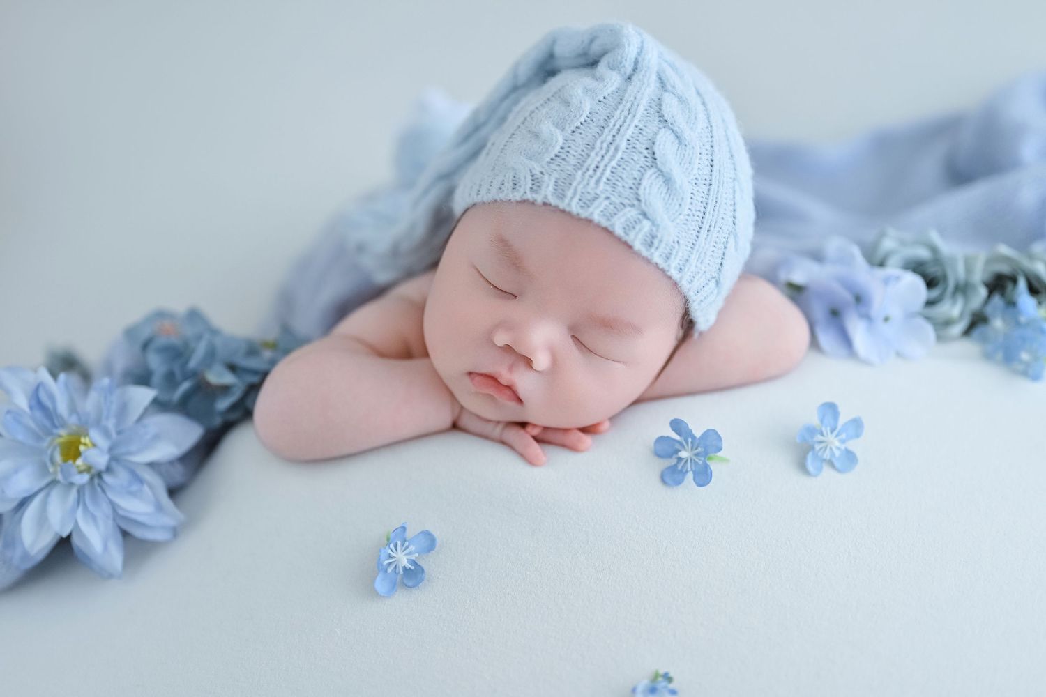 Peaceful newborn baby in gray knit hat resting chin on hands among blue flowers on white backdrop.