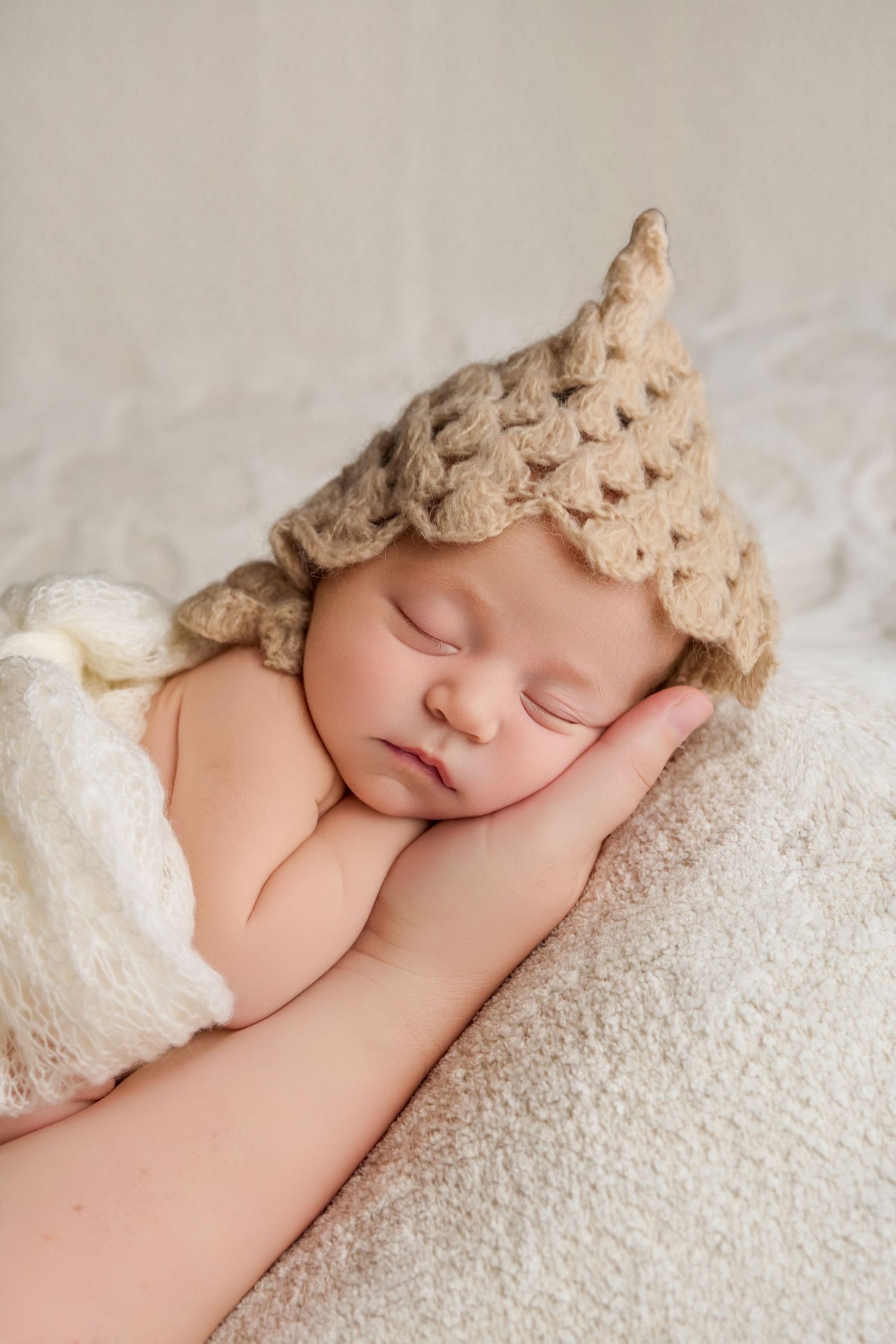 Sleeping newborn baby wearing a beige crochet crown hat, resting peacefully on a soft cream blanket.