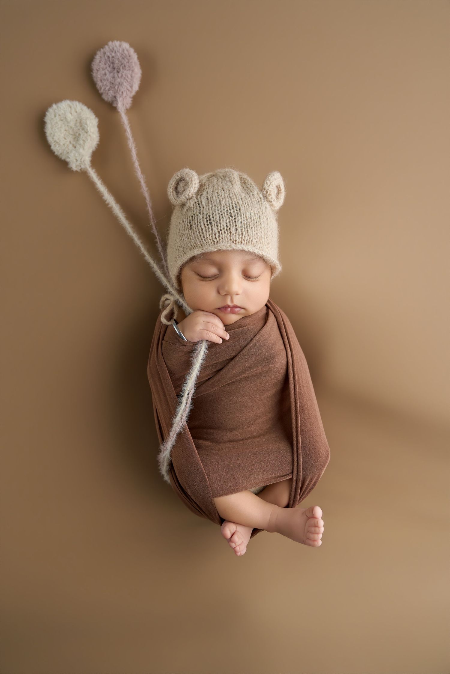 Sleeping newborn baby wrapped in brown swaddle wearing a knit bear ear hat, holding decorative balloon stems on beige background.