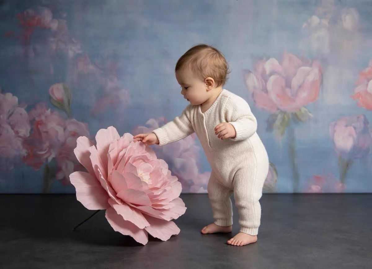 Baby in white romper touching a large pink peony flower against a floral painted backdrop.