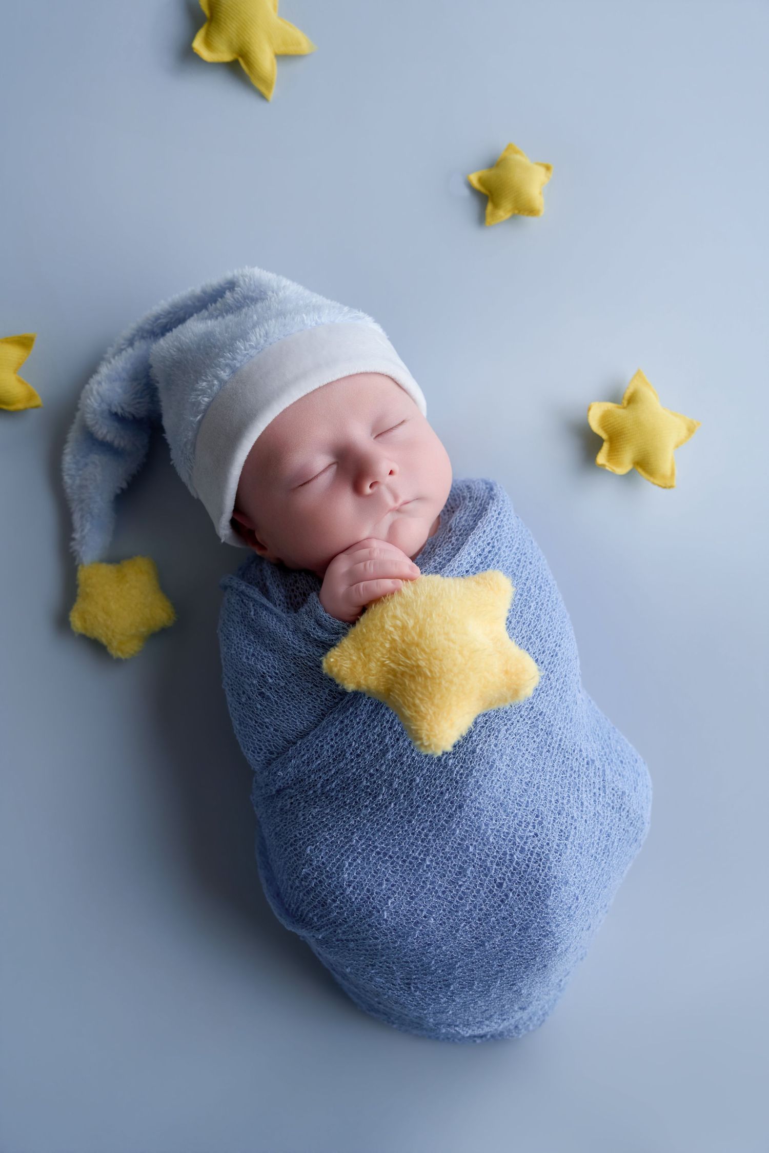 Newborn baby swaddled in blue wrap wearing a nightcap, sleeping peacefully surrounded by yellow felt stars.