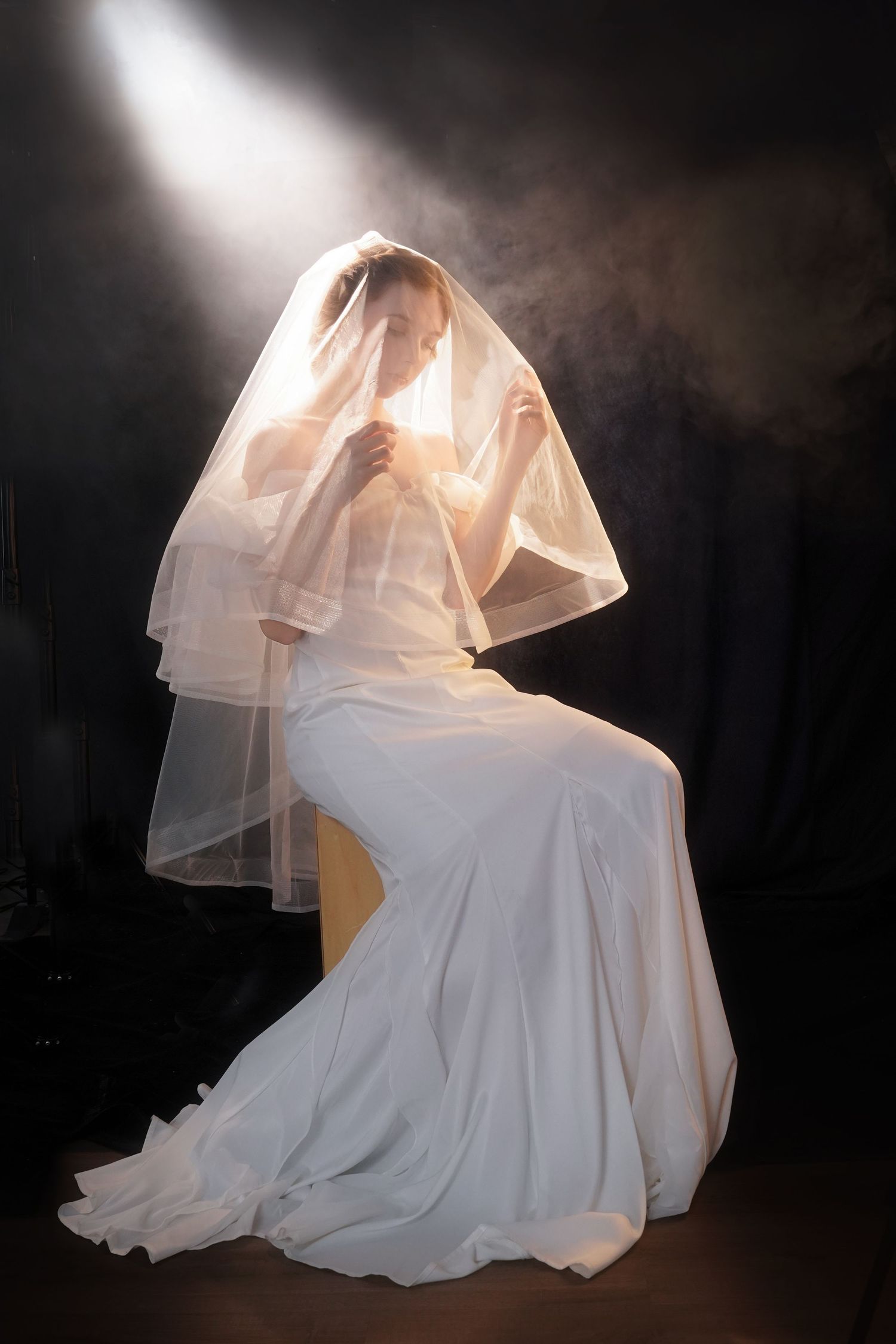 Bride in white wedding gown and veil sits dramatically illuminated by spotlight against dark smoky background.