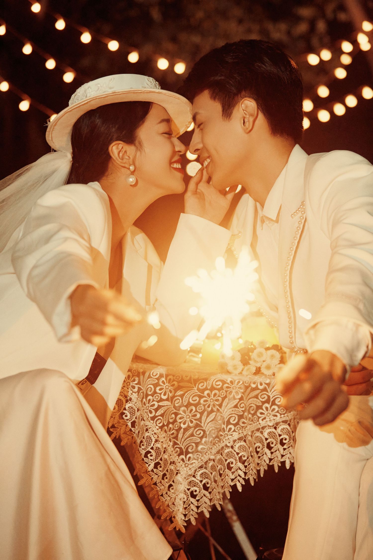 A couple in white wedding attire share a romantic moment holding sparklers under string lights.