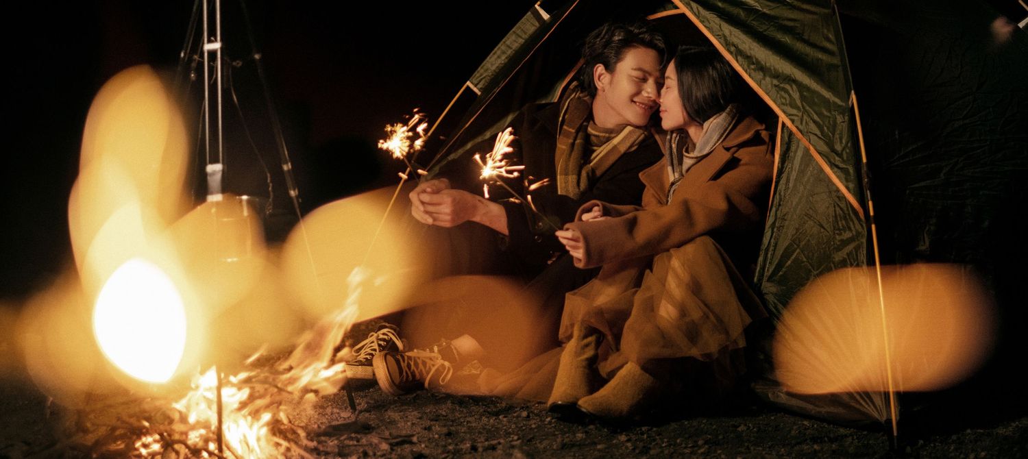 Couple cuddling by a campfire outside a tent at night with warm glowing bokeh lights.