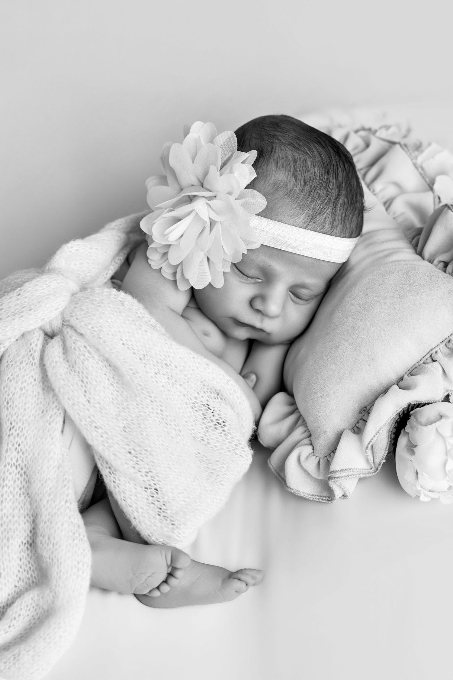 Black and white photo of a sleeping newborn baby girl wearing a floral headband, wrapped in a soft blanket.
