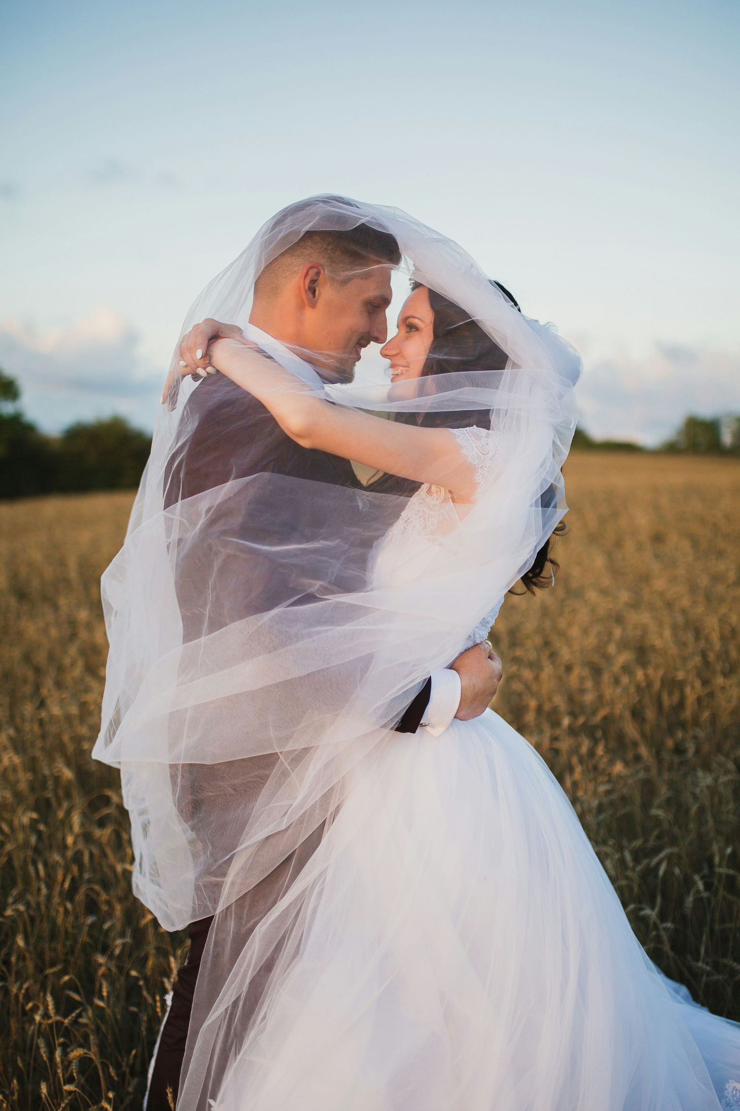Bride and groom embracing under flowing veil in golden wheat field at sunset.