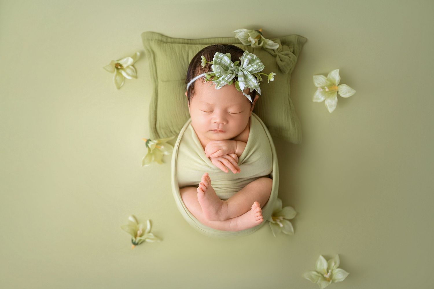 Newborn baby girl sleeping peacefully in green bowl with floral headband and white flowers on green background.