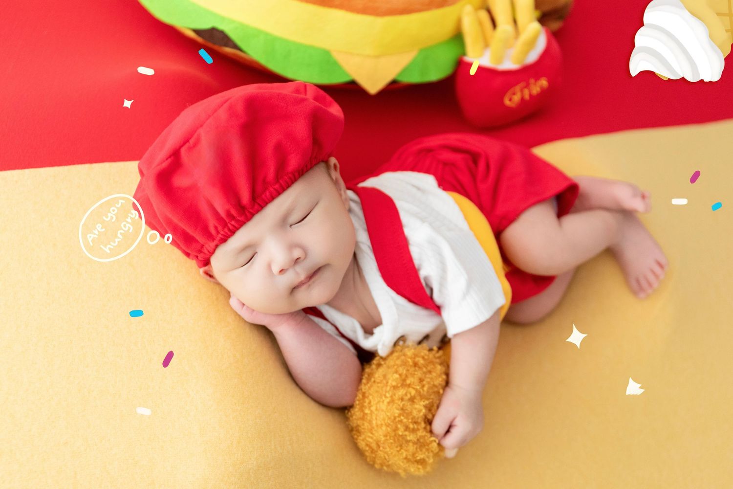 Sleeping baby dressed in a fast food worker costume holding a fried chicken piece on a red and yellow background.