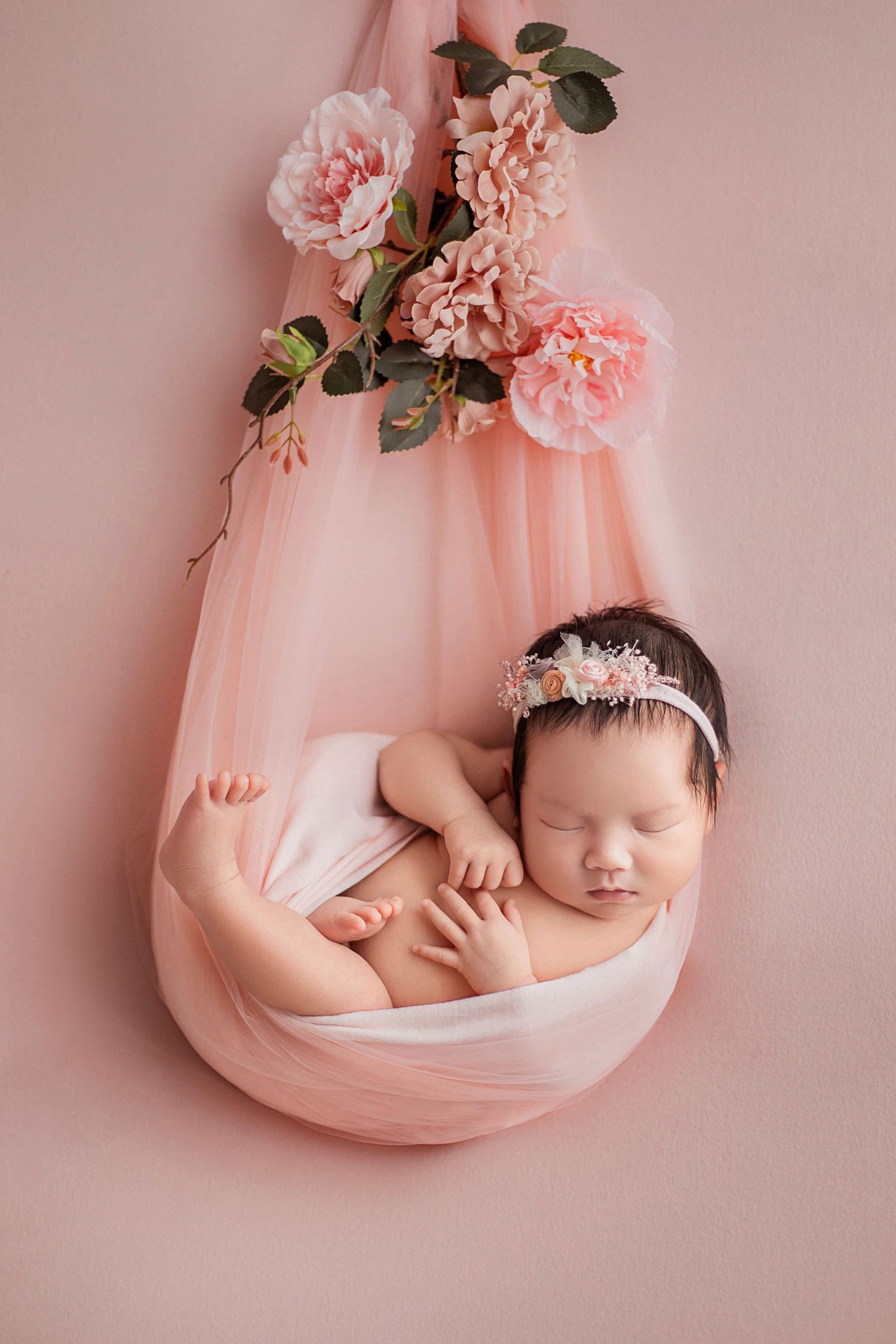 Peaceful newborn baby girl in floral headband rests in pink tulle sling adorned with blush peonies.