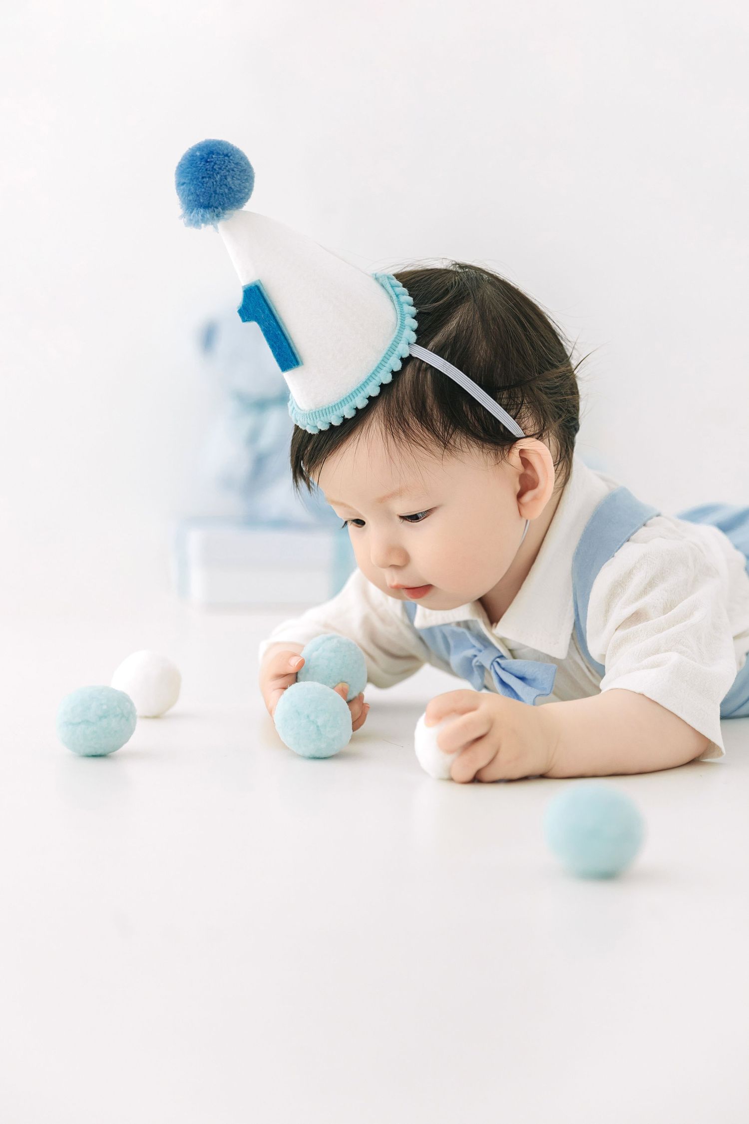 Cute baby wearing a blue number one birthday hat playing with colorful pom pom balls on white floor.