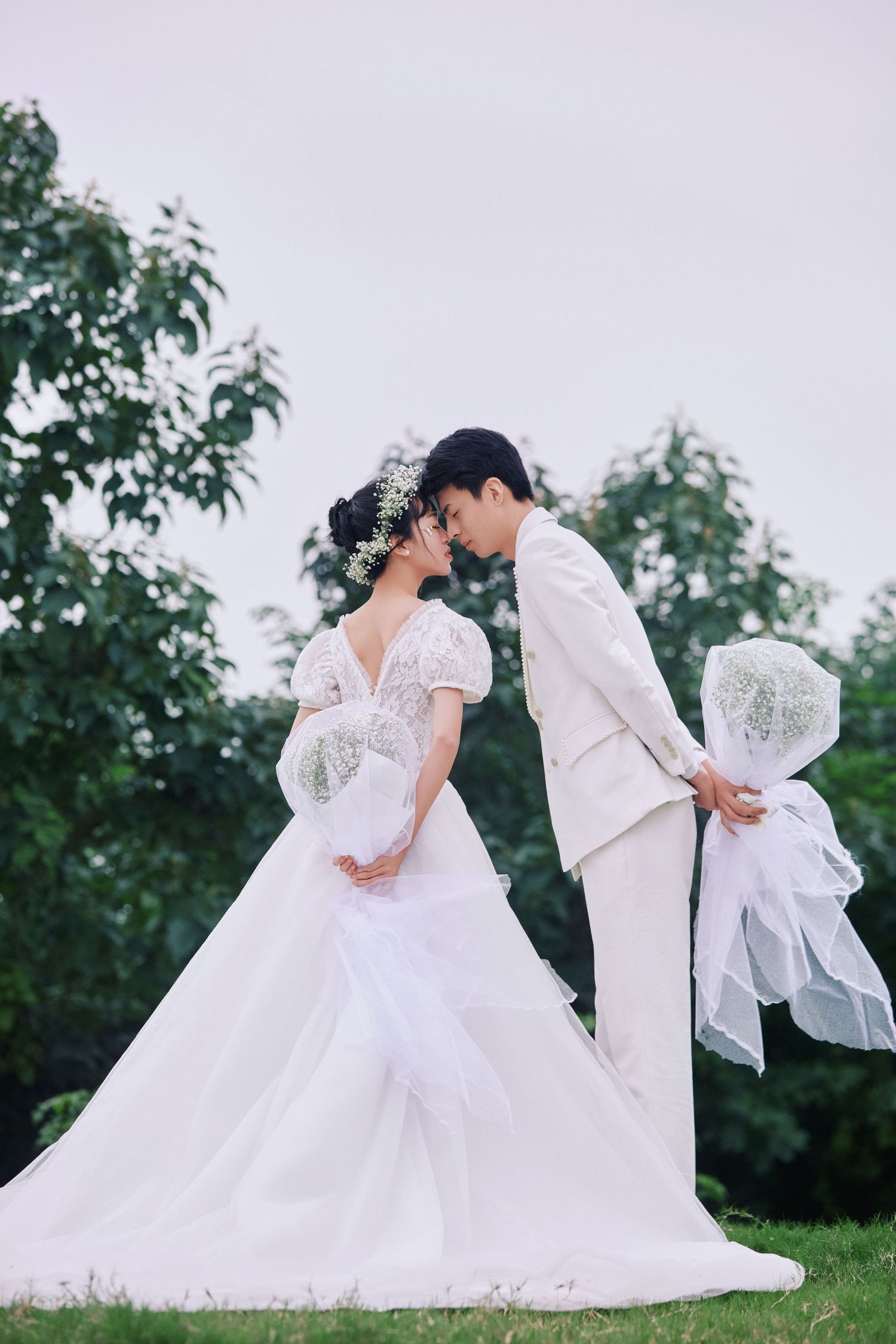 Romantic couple in white wedding attire sharing an intimate moment outdoors with lush green trees in background.
