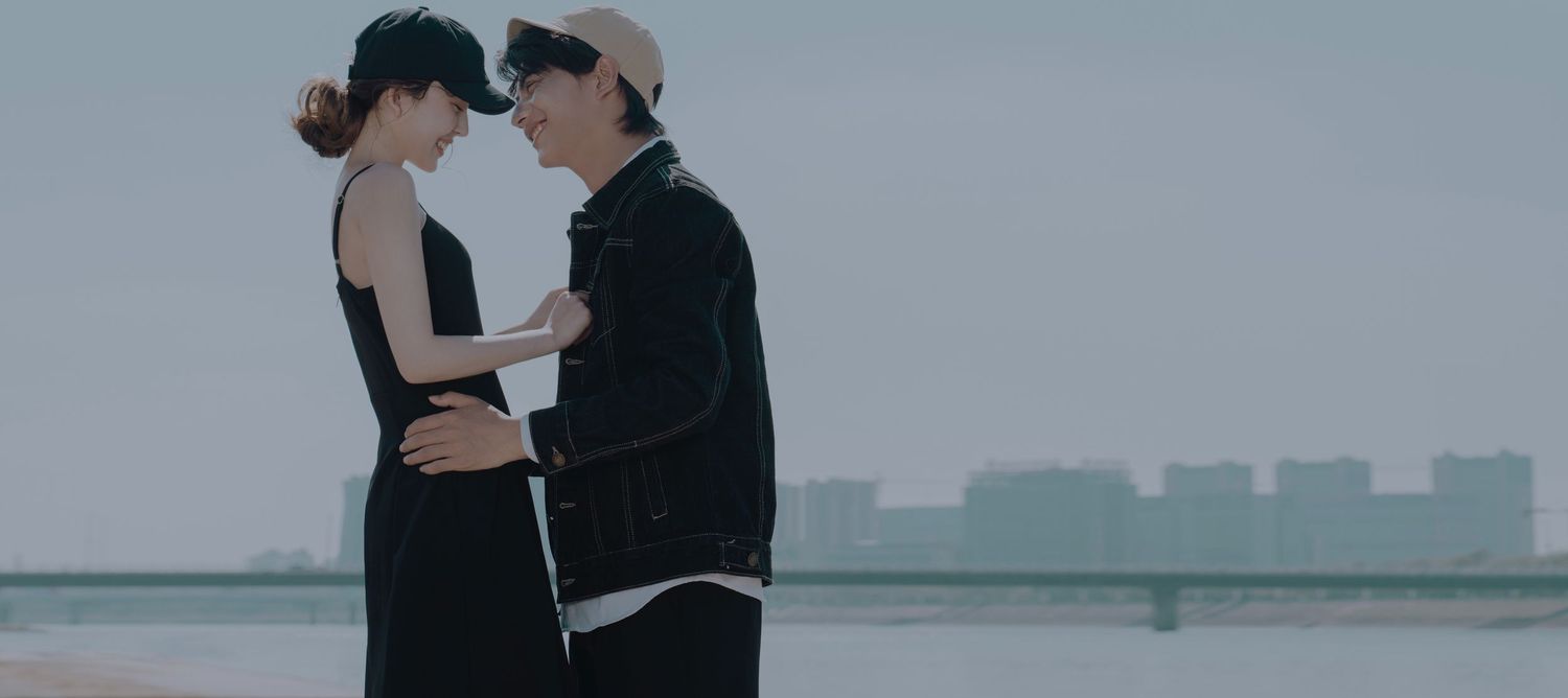 A young couple in black outfits standing close together in an intimate moment by a waterfront.