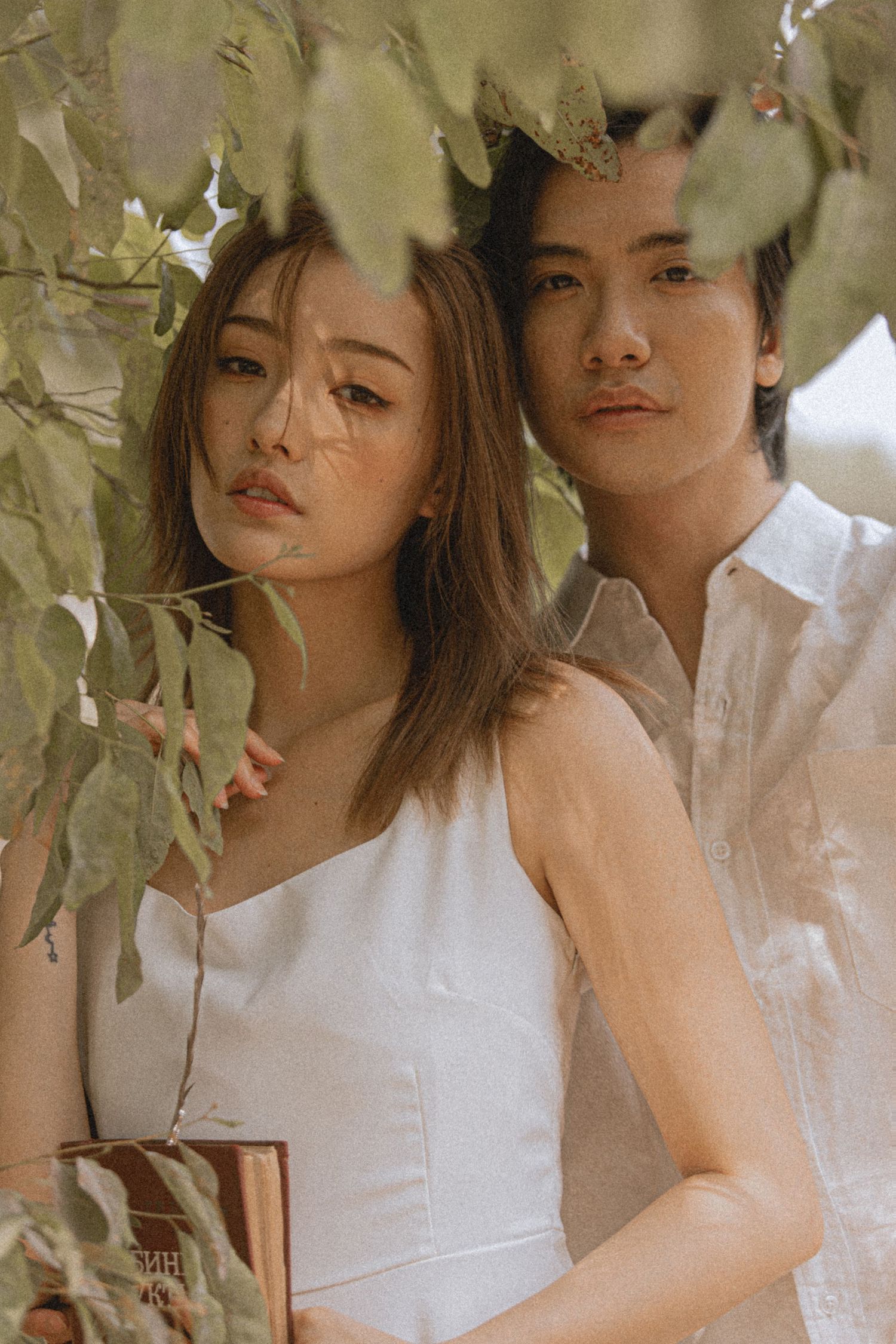 Young Asian couple in white attire pose romantically under green leaves in a soft, natural light outdoor setting.