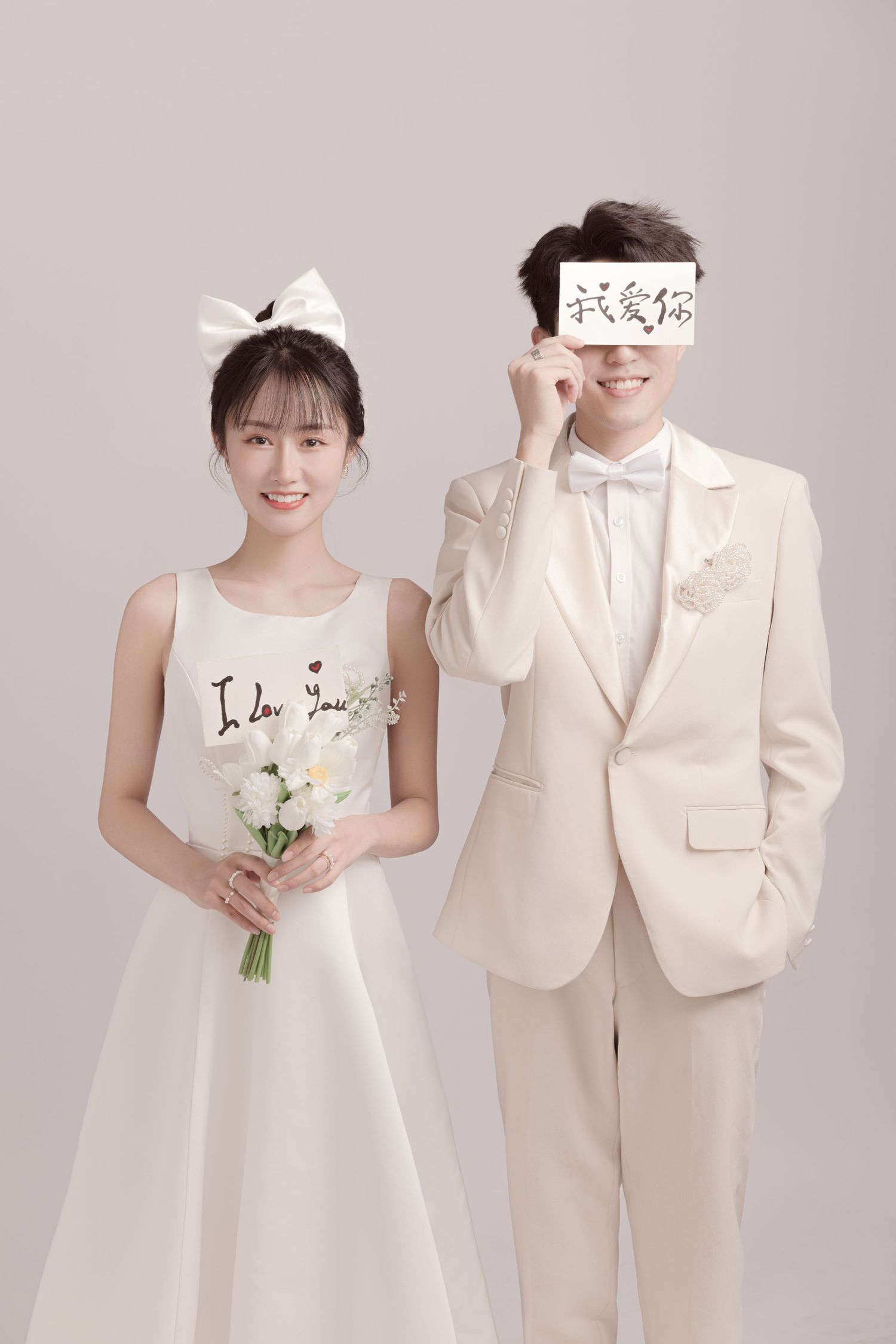 Bride in white dress with bow headband and groom in cream suit holding an I love you sign indoors.