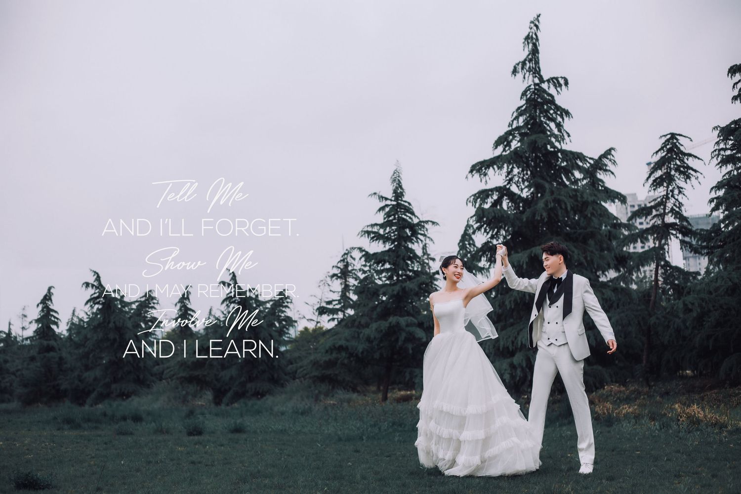 Bride and groom dance joyfully on green lawn surrounded by tall evergreen trees on cloudy day.