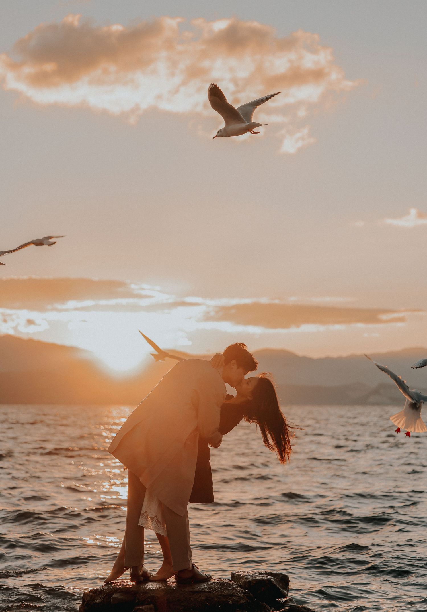 A couple shares a romantic kiss on rocks by the sea at sunset surrounded by flying seagulls.
