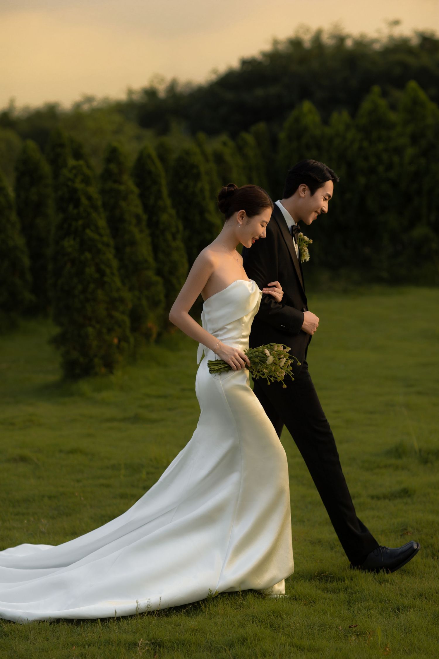 Elegant bride and groom walk together at sunset on a green lawn lined with tall cypress trees.