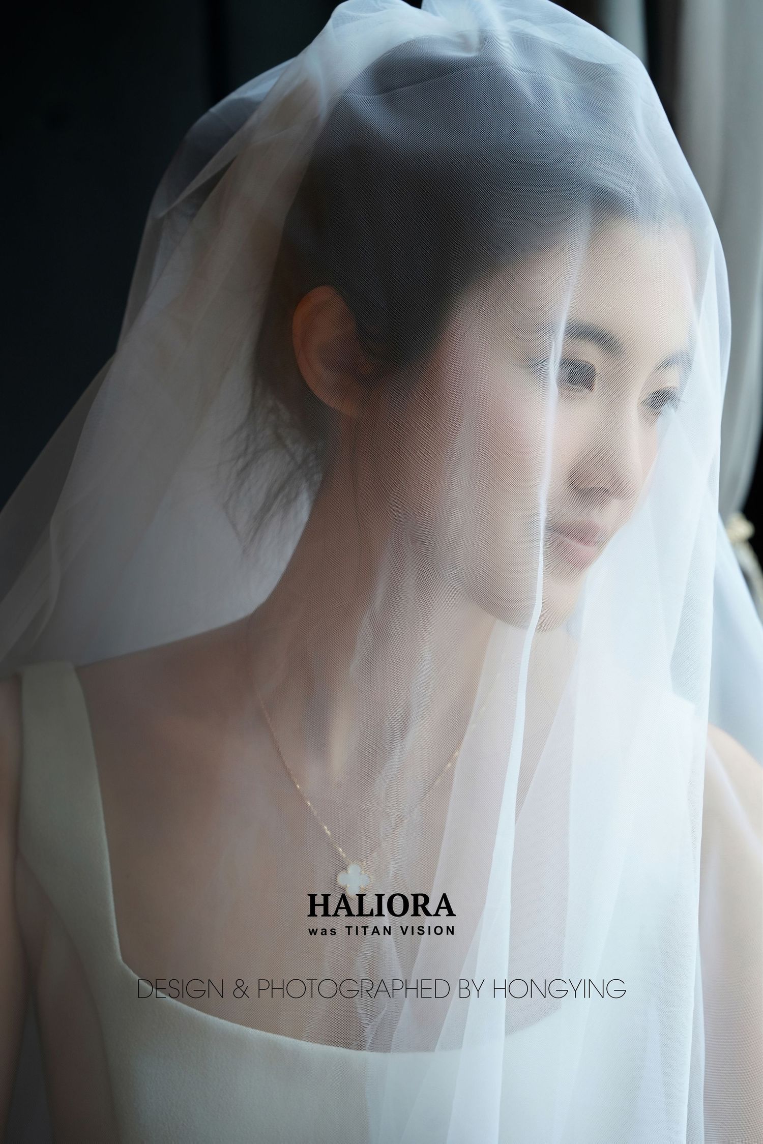 Elegant Asian bride draped in a sheer white veil gazes softly downward, wearing a delicate necklace indoors.