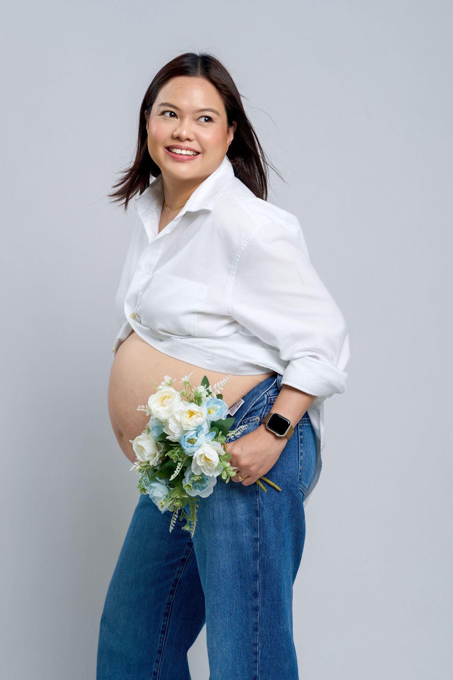 Smiling pregnant Asian woman in white shirt and jeans holding blue and white flowers, posing against gray background.