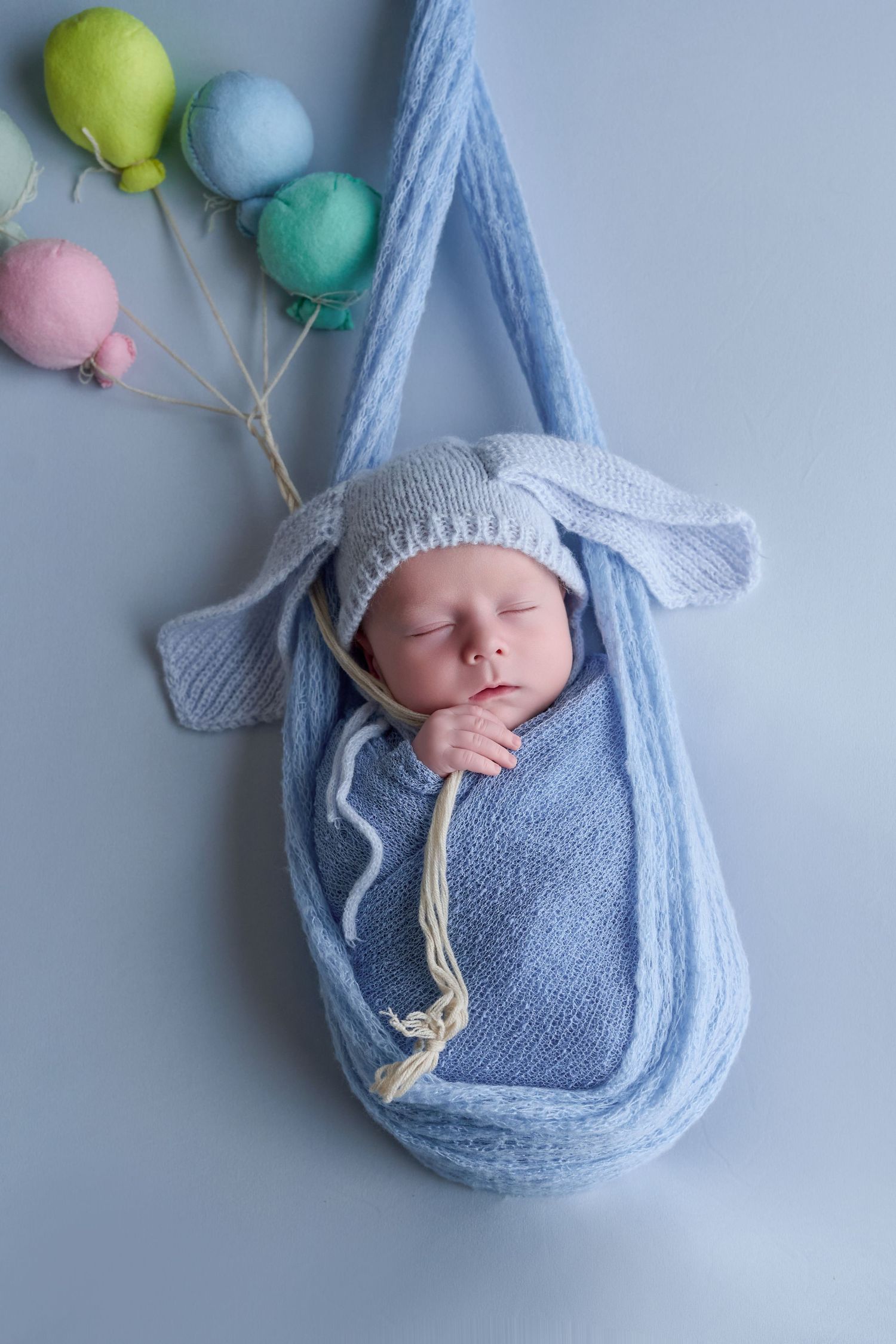 Sleeping newborn baby wrapped in blue knit swaddle wearing bunny ear hat, lying in hanging cradle with colorful balloon decorations.