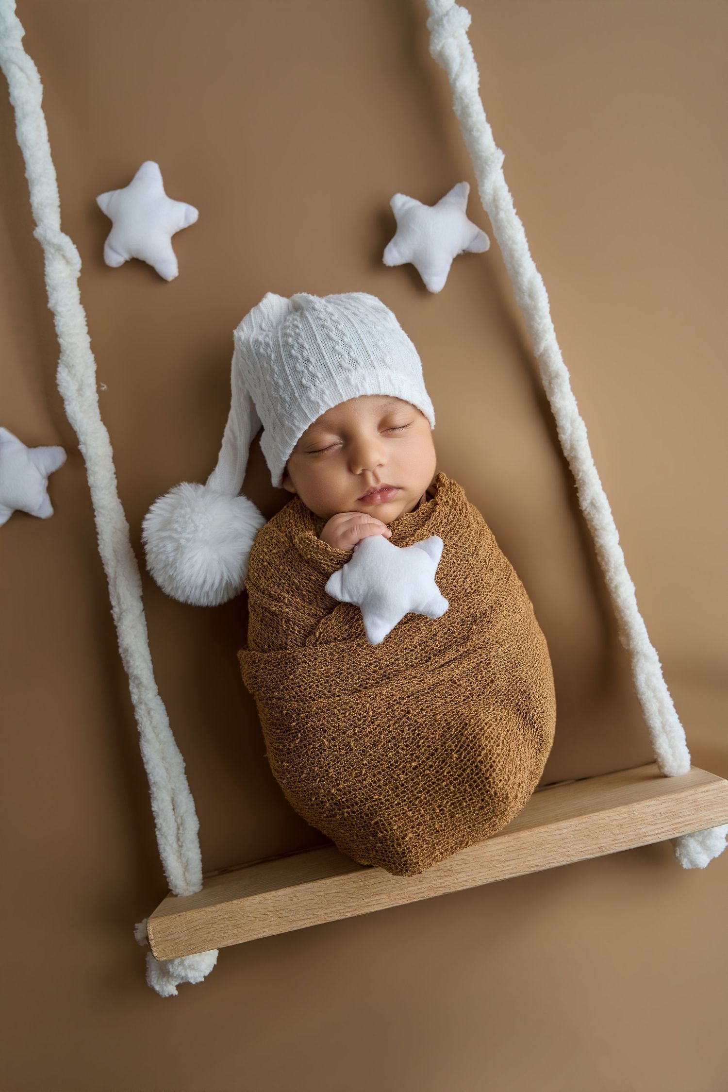 Newborn baby swaddled in brown wrap wearing white knit hat, sleeping on wooden swing with white star decorations.