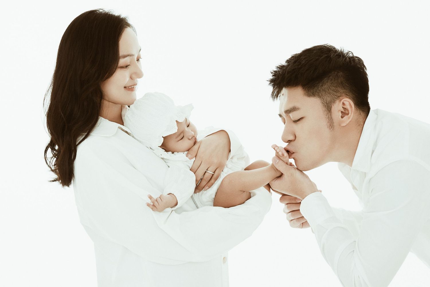 Tender moment as Asian father kisses baby's hand while mother holds newborn in white studio setting.
