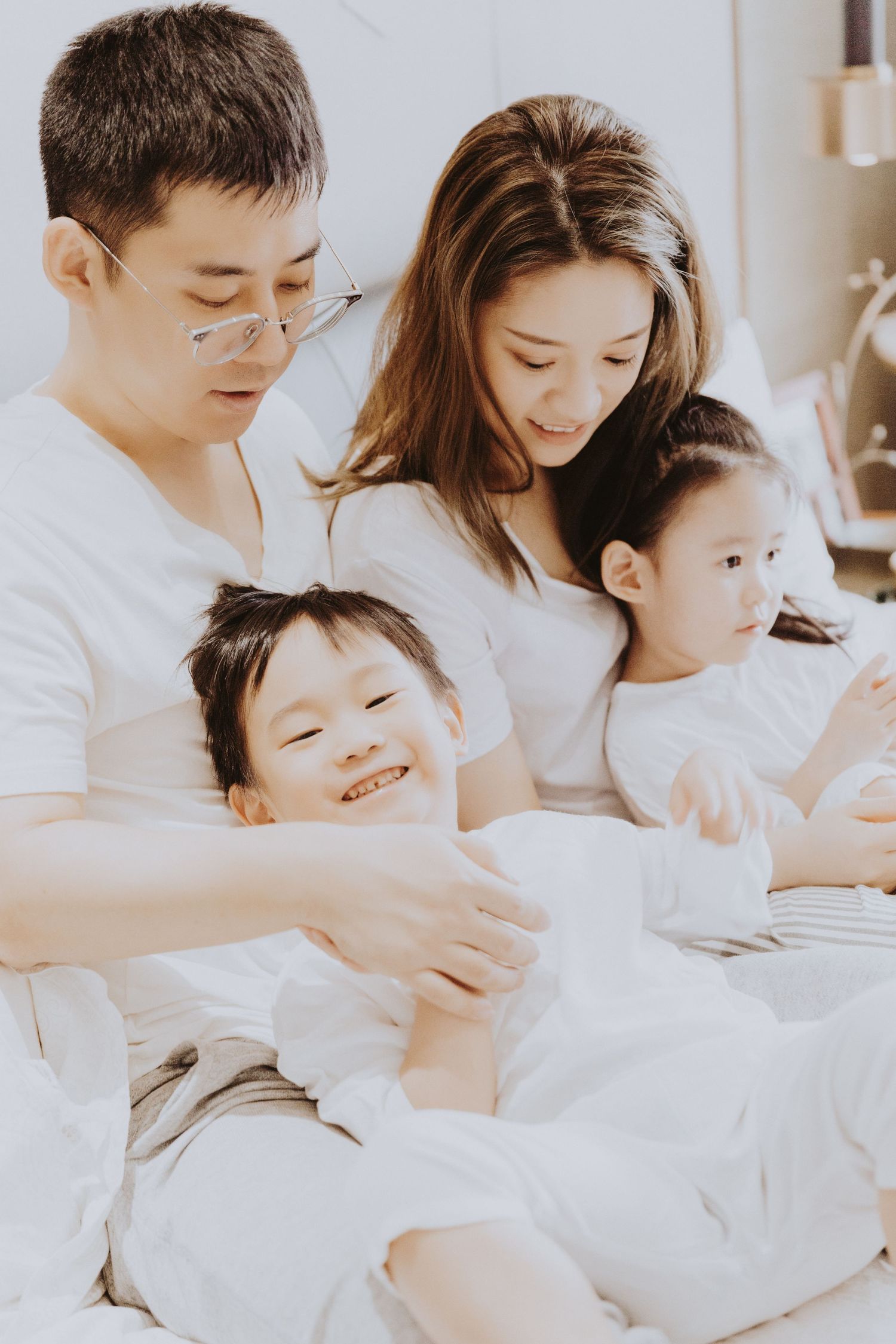 An Asian family shares a tender moment on a white bed, with a father tickling a laughing young boy.