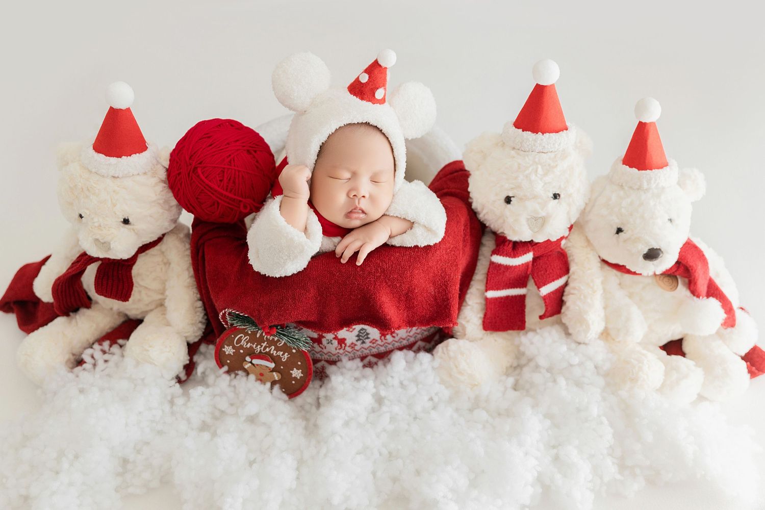 Newborn baby in Santa hat and white costume lying with white teddy bears in Christmas outfits on snowy surface.