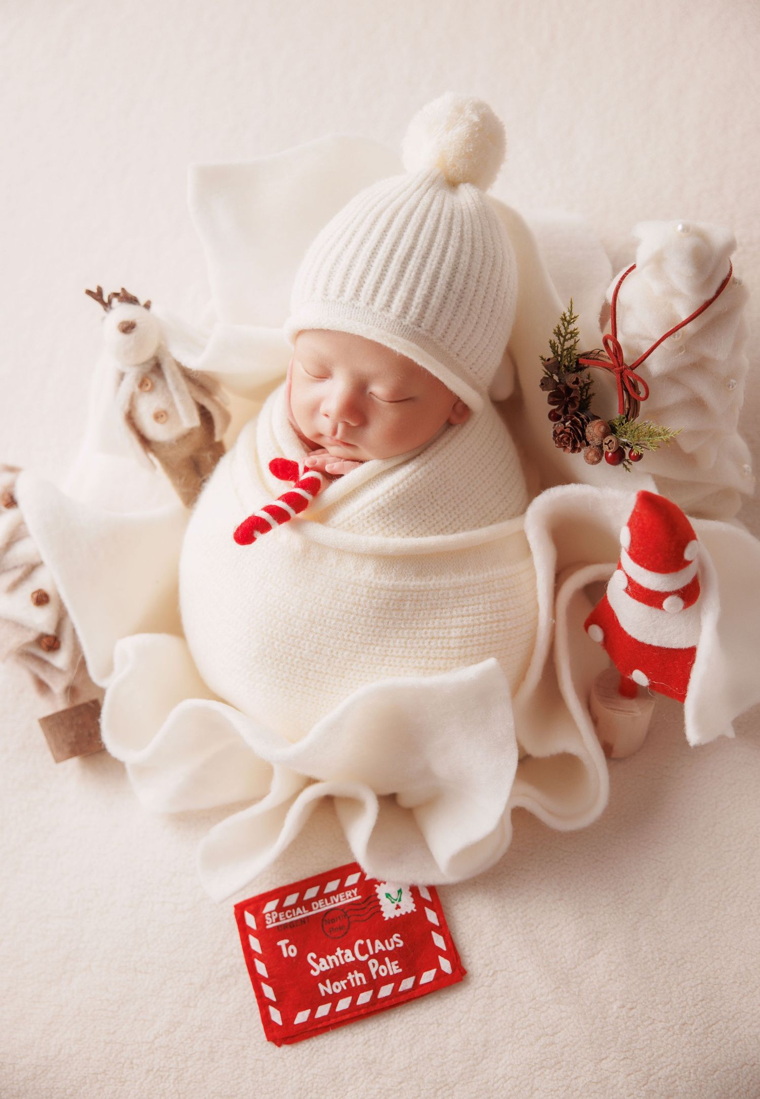 Sleeping newborn wrapped in cream swaddle with white pom-pom hat beside a Santa Claus North Pole letter.