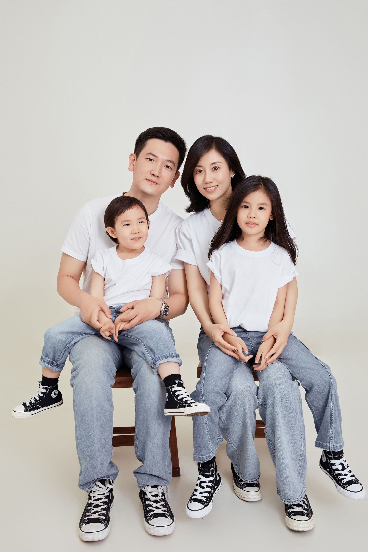 Asian family of four in white tees and jeans sit together on a bench in a bright studio portrait.